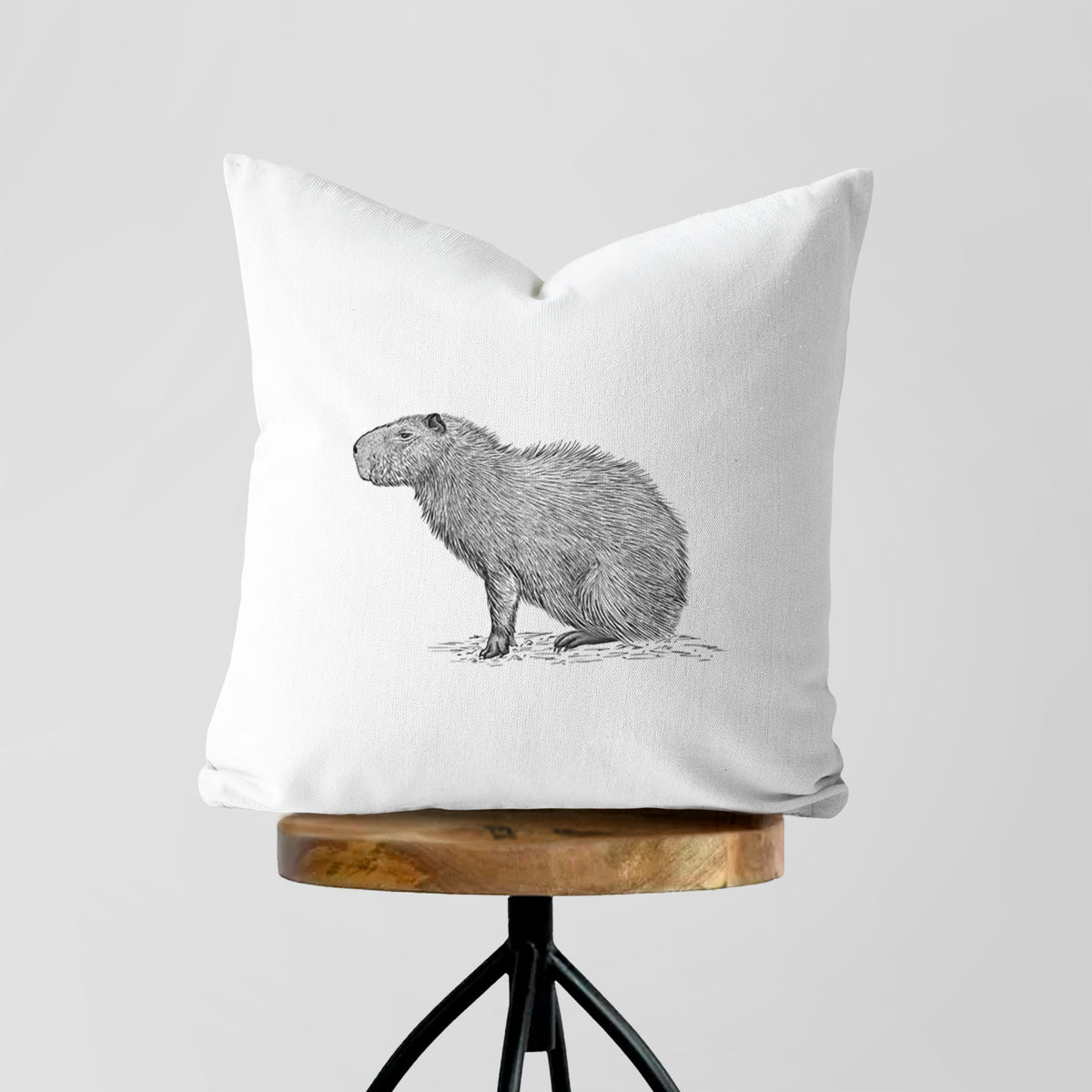 White pillow cover printed with a detailed black ink illustration of a capybara standing in profile, on a wooden stool.