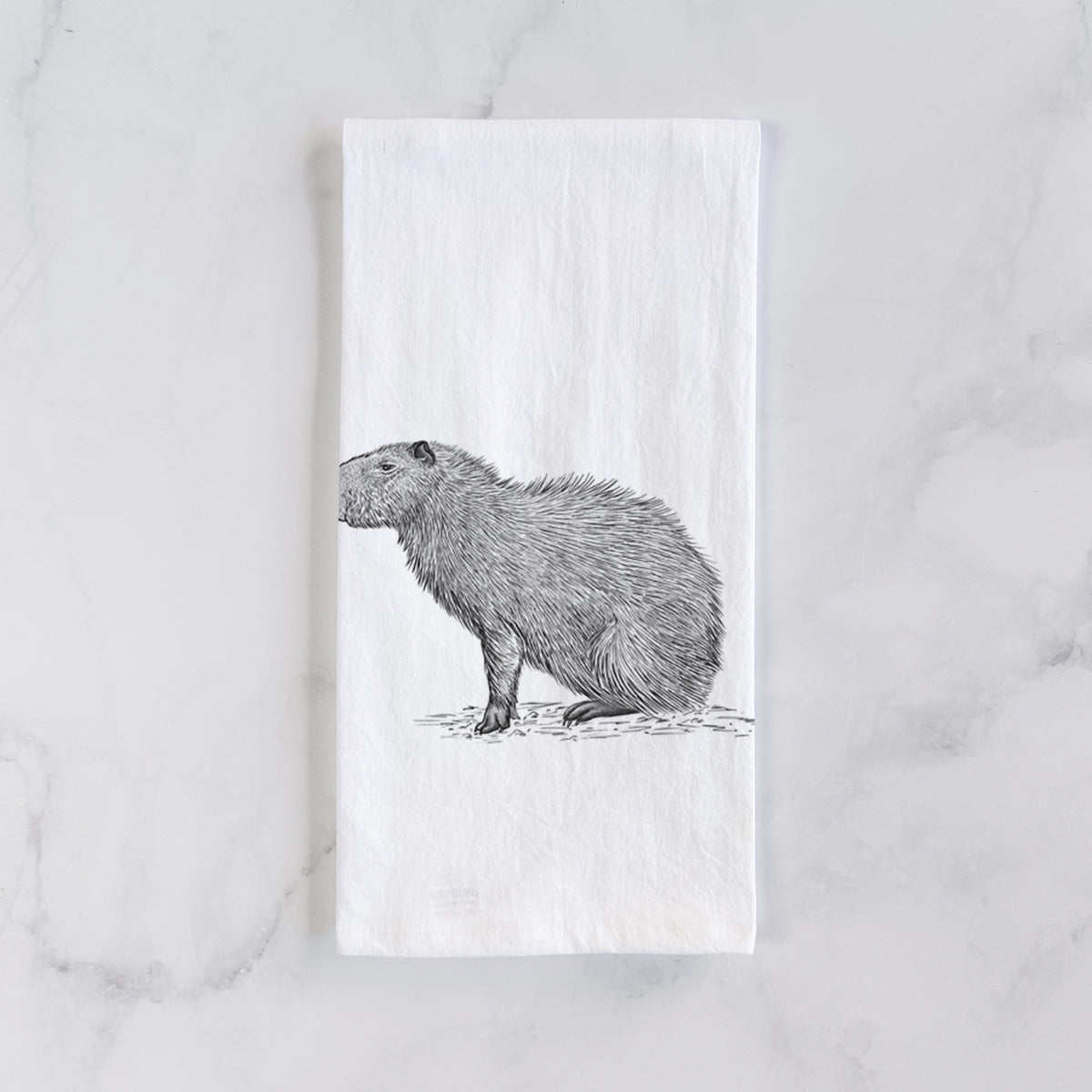 White cotton tea towel laid flat on marble, printed with a detailed black ink illustration of a capybara standing in profile.