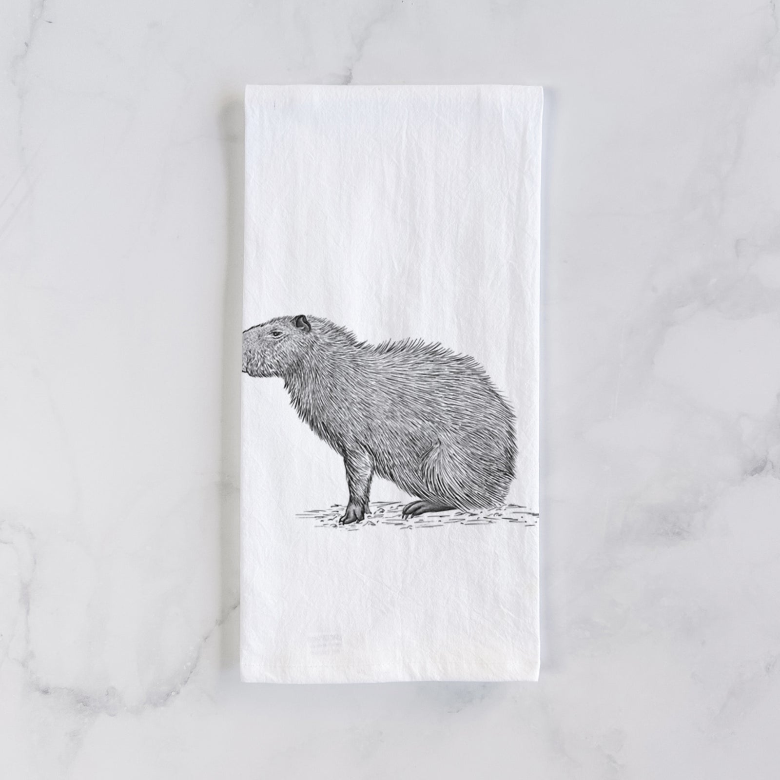 White cotton tea towel printed with a detailed black ink illustration of a capybara standing in profile, on a marble counter beside a round wooden cutting board and greenery.