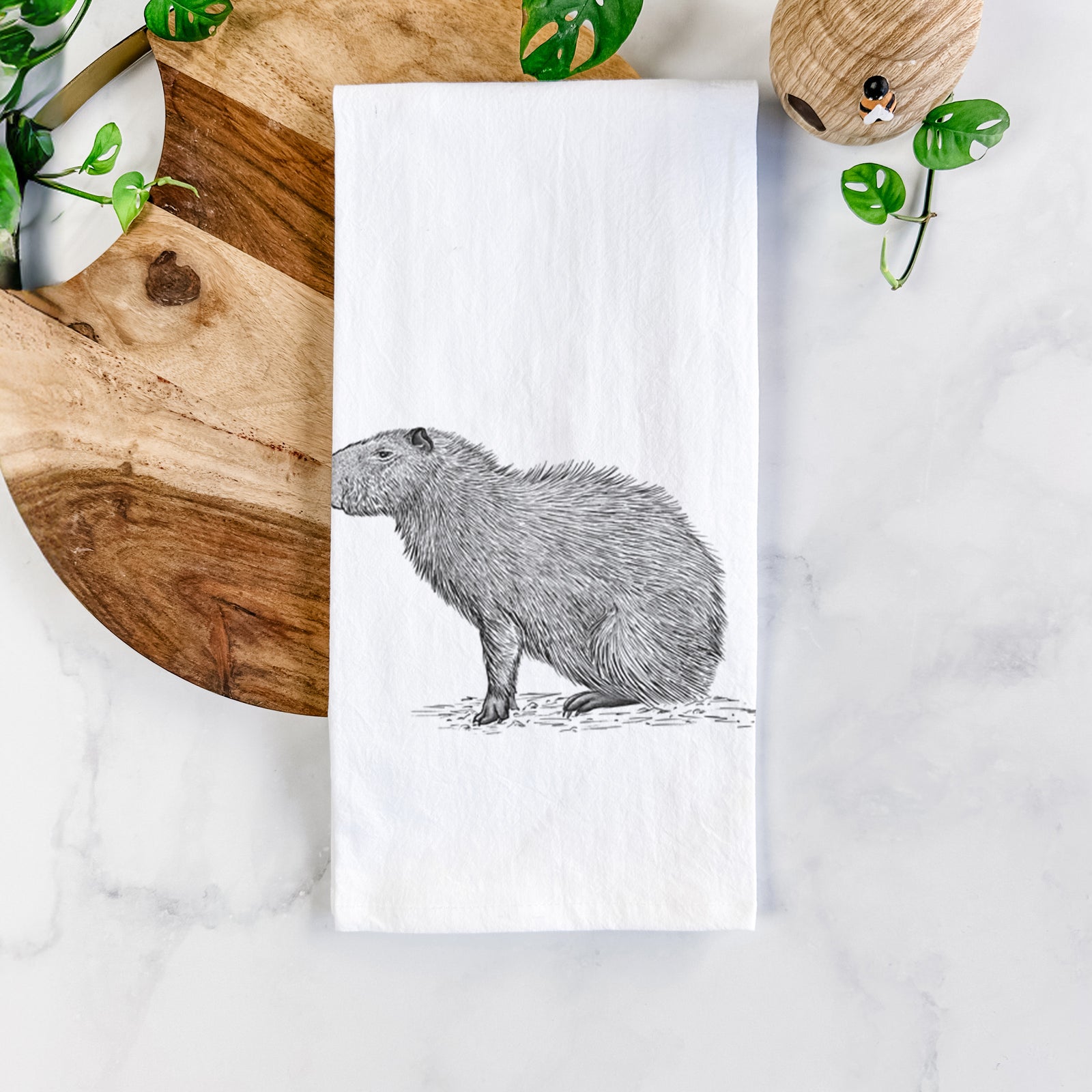 White cotton tea towel printed with a detailed black ink illustration of a capybara standing in profile, on a marble counter beside a round wooden cutting board and greenery.