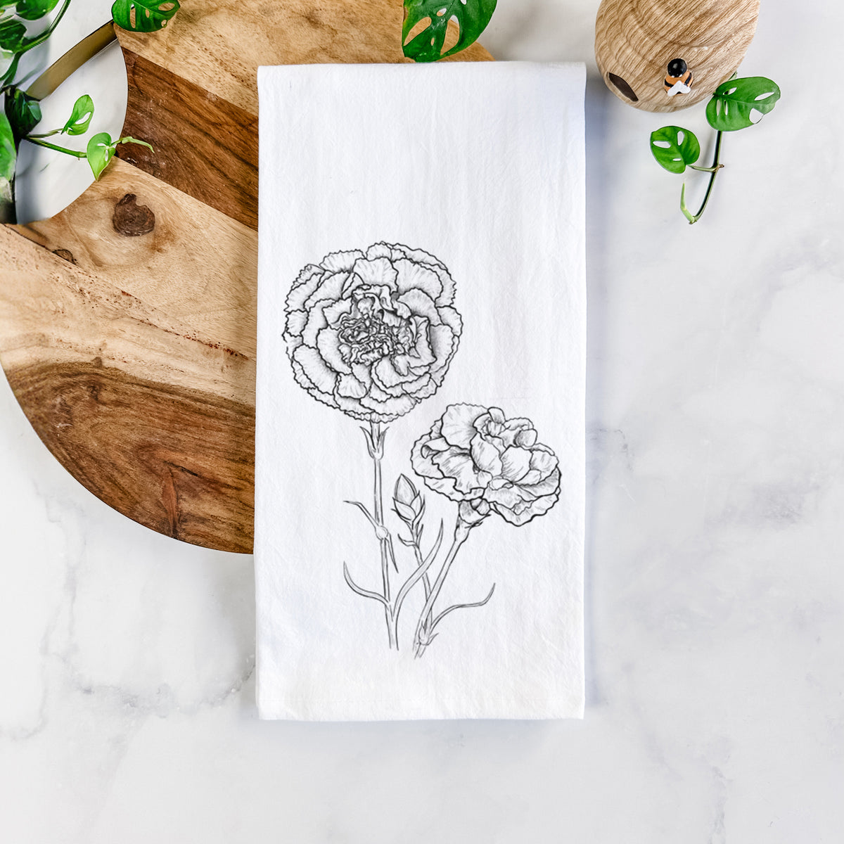 White cotton tea towel printed with a black ink line drawing of carnation flowers on stems, on a marble counter beside a round wooden cutting board and greenery.