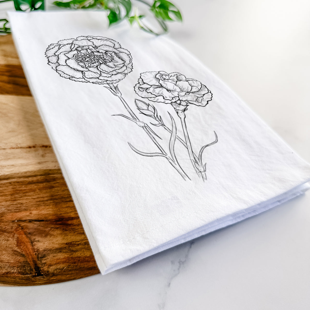 Close-up of a folded white tea towel on a wooden board, showing a black ink line drawing of carnation flowers on stems.