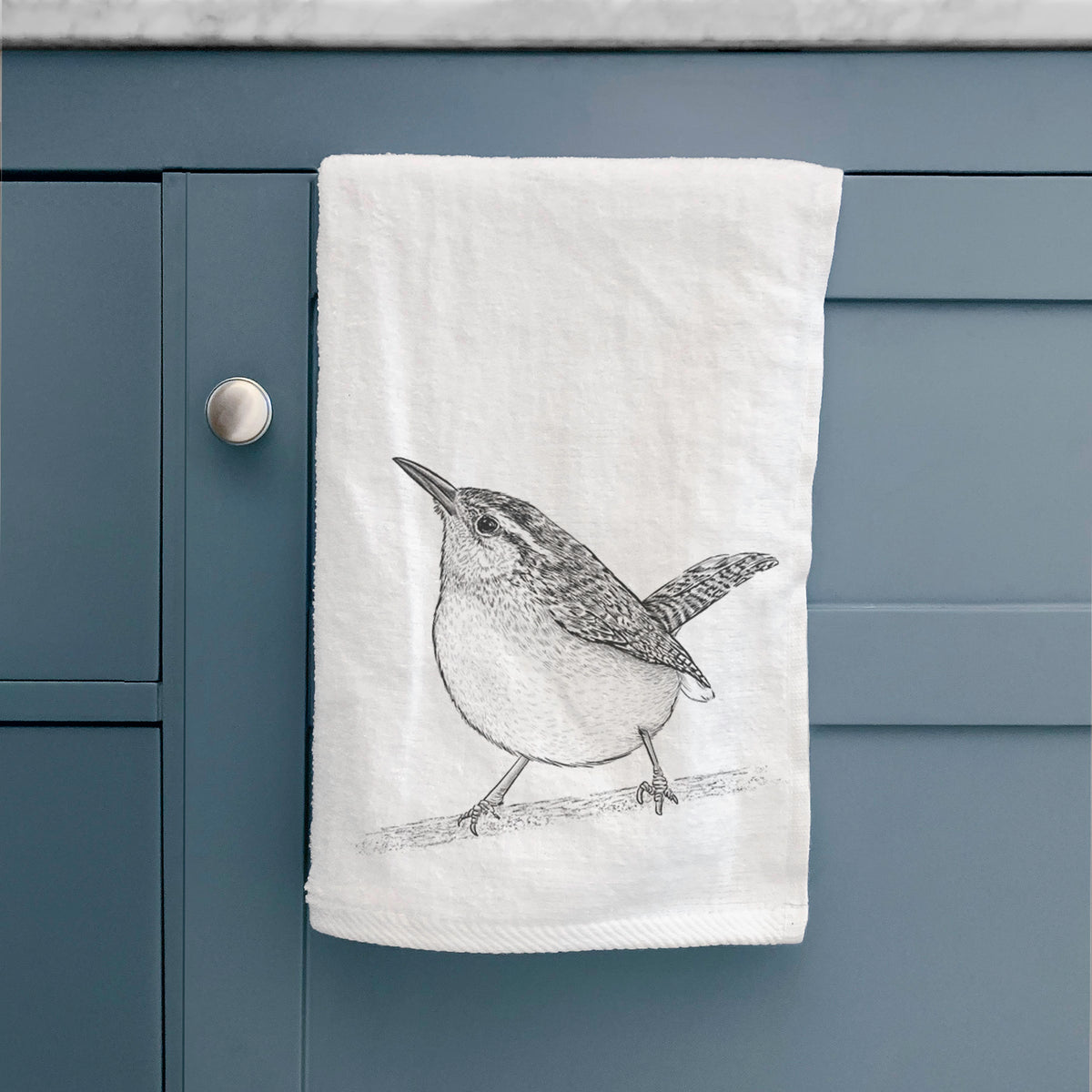 White hand towel printed with a detailed black ink sketch of a Carolina wren perched on a branch with its tail raised, hanging on a cabinet door.