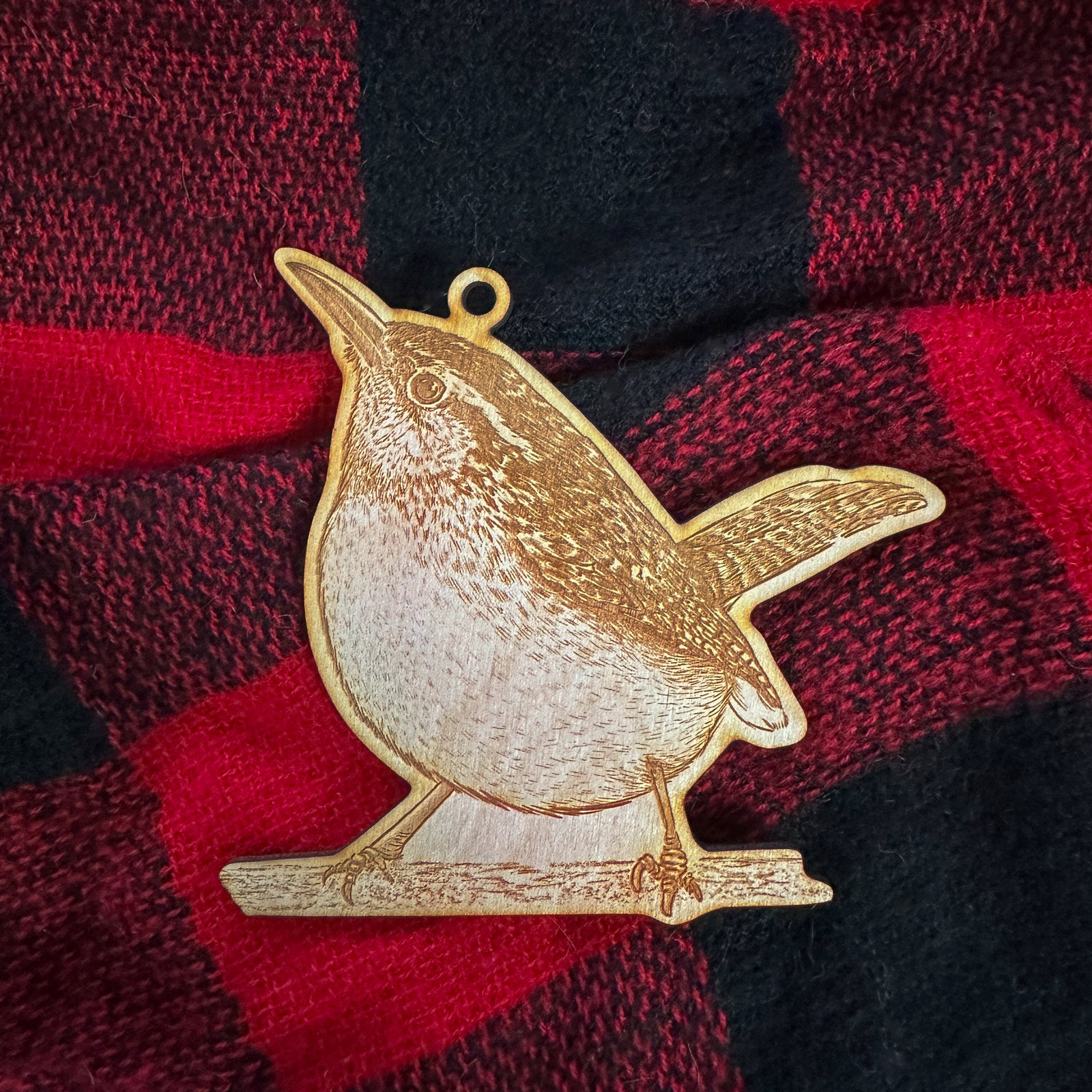 Wooden ornament featuring a detailed engraving of a Carolina wren perched on a branch with its tail raised, on a knit blanket with pine cones.