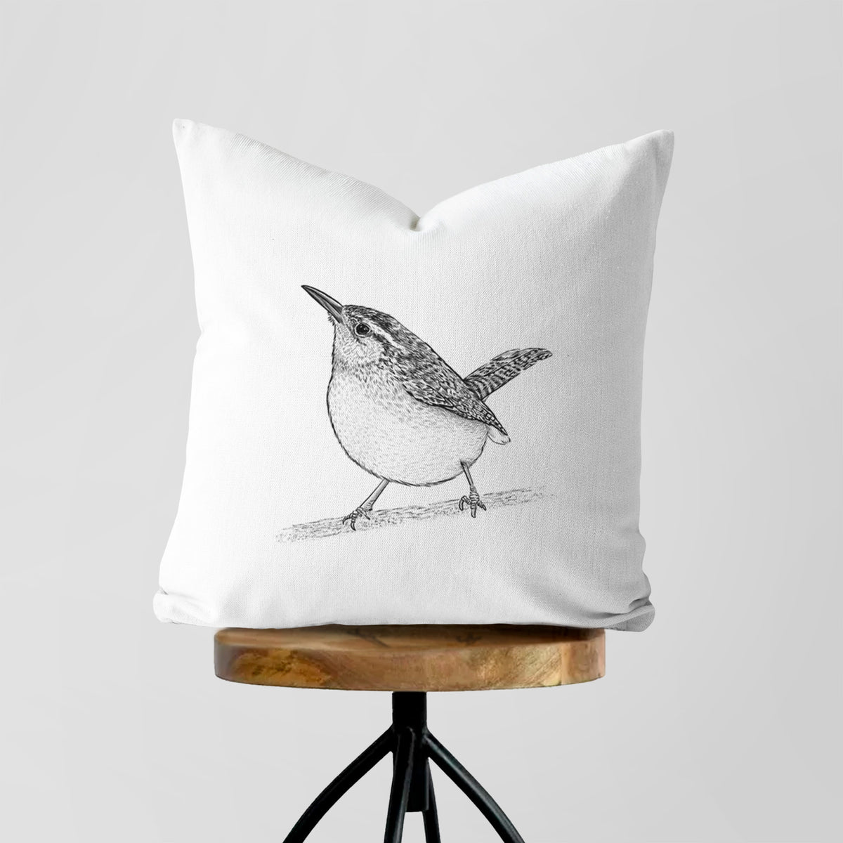 White pillow cover printed with a detailed black ink sketch of a Carolina wren perched on a branch with its tail raised, on a wooden stool.