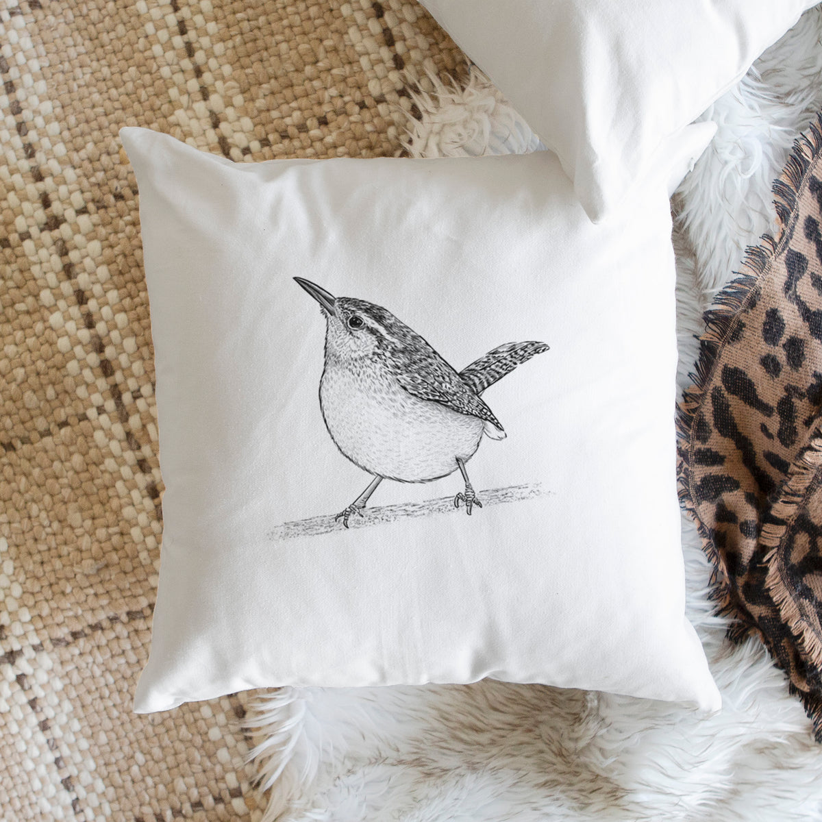 White pillow cover printed with a detailed black ink sketch of a Carolina wren perched on a branch with its tail raised, on a woven chair.