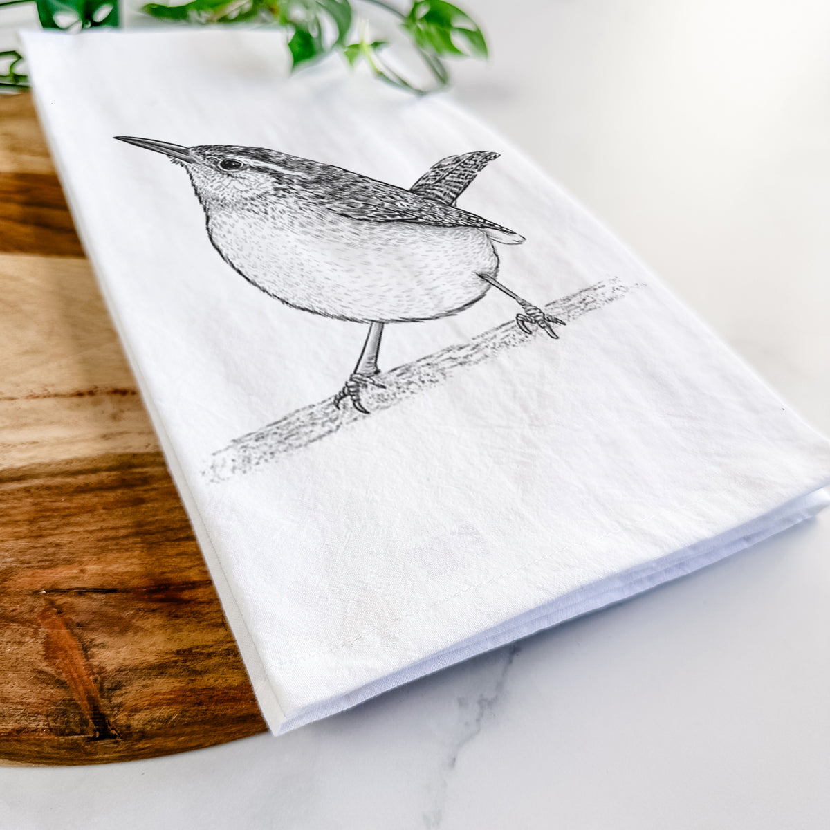 Close-up of a folded white tea towel on a wooden board, showing a detailed black ink sketch of a Carolina wren perched on a branch with its tail raised.