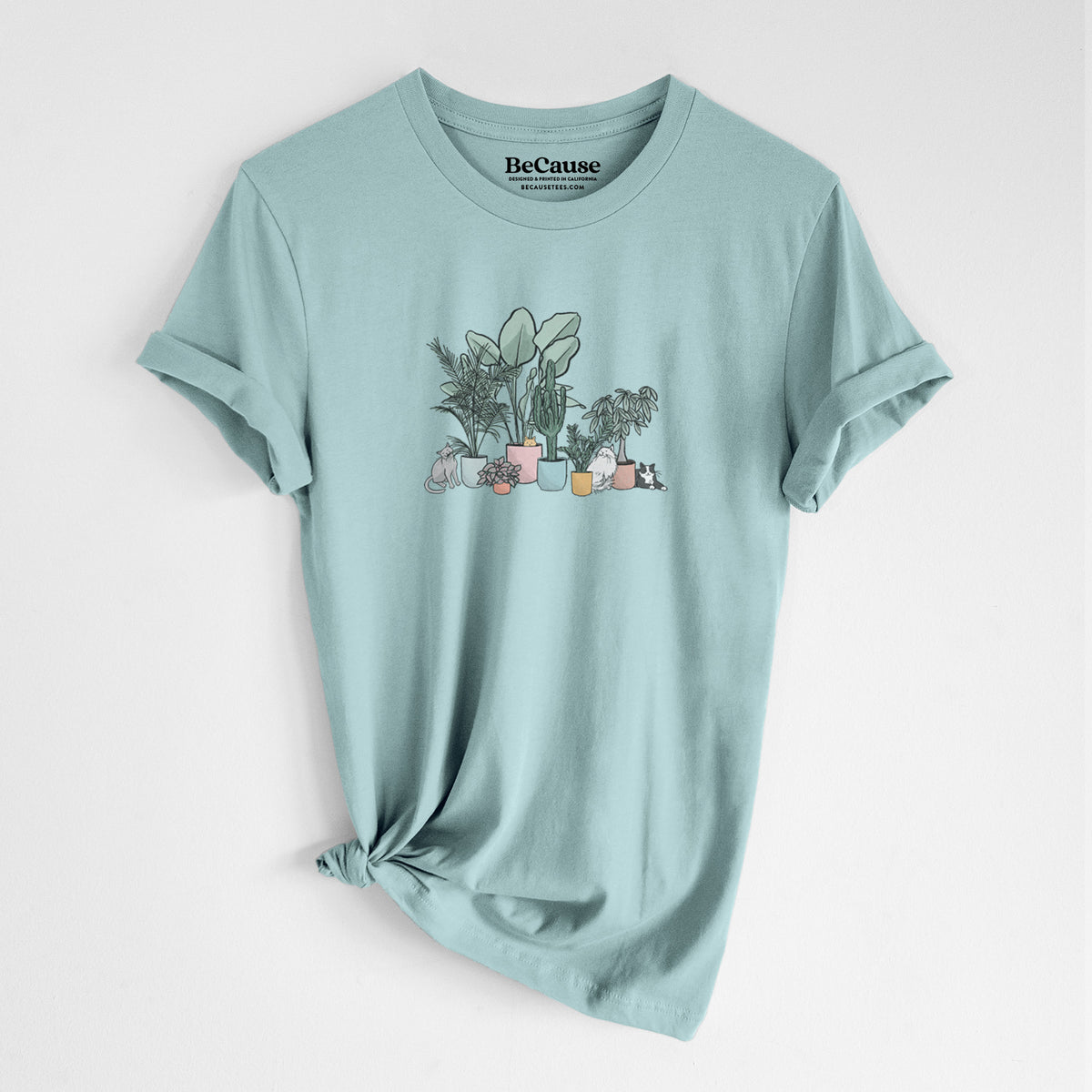 Dusty blue lightweight cotton t-shirt laid flat with the sleeves rolled, printed with a full-color illustration of cats lounging among potted houseplants.