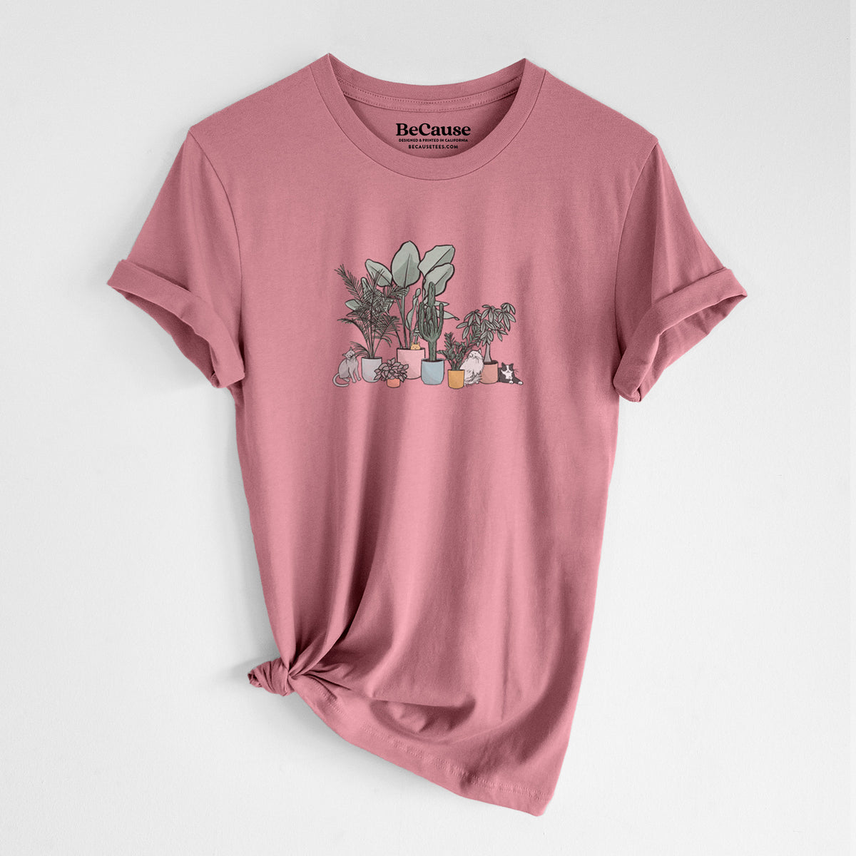 Mauve pink lightweight cotton t-shirt laid flat with the sleeves rolled, printed with a full-color illustration of cats lounging among potted houseplants.