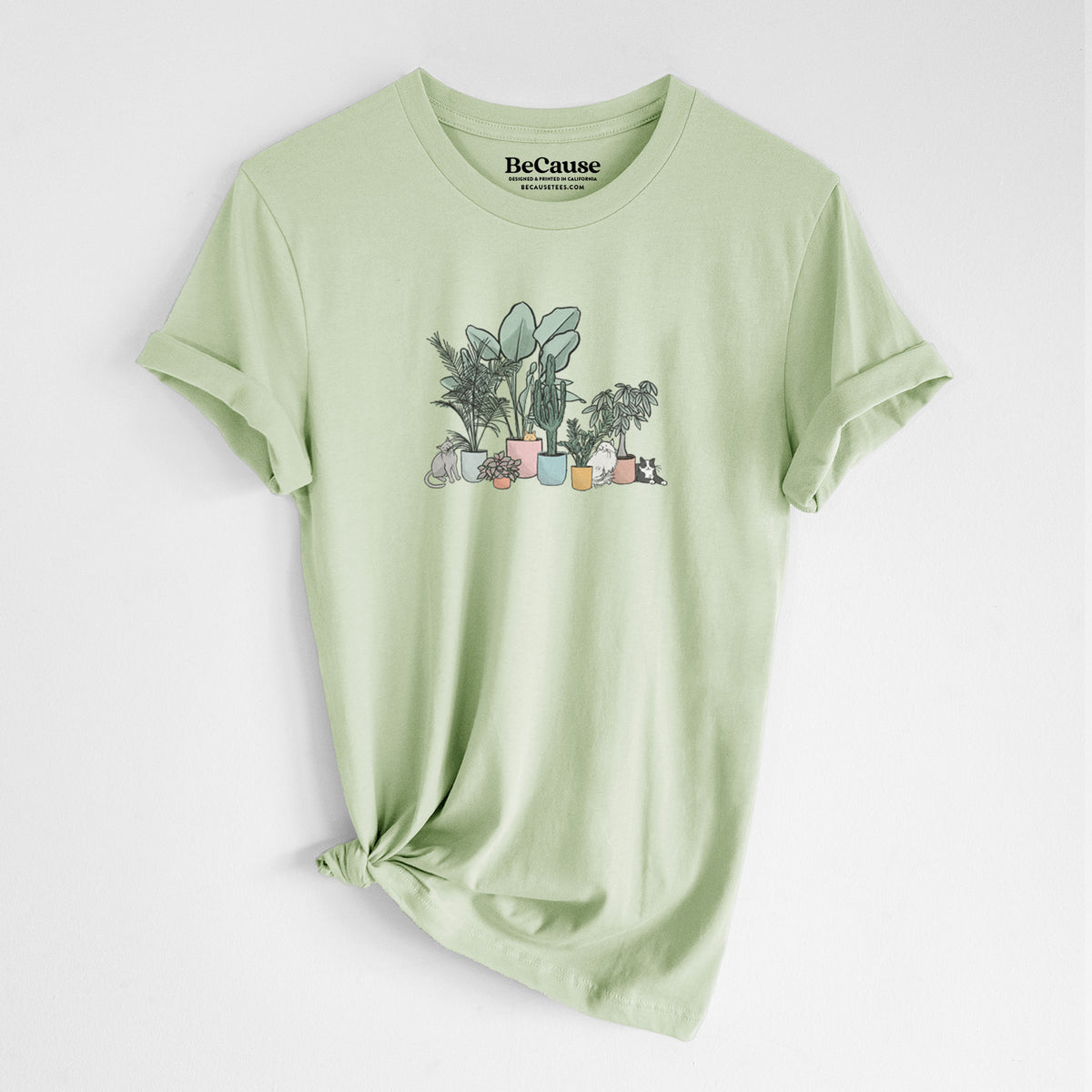 Spring green lightweight cotton t-shirt laid flat with the sleeves rolled, printed with a full-color illustration of cats lounging among potted houseplants.