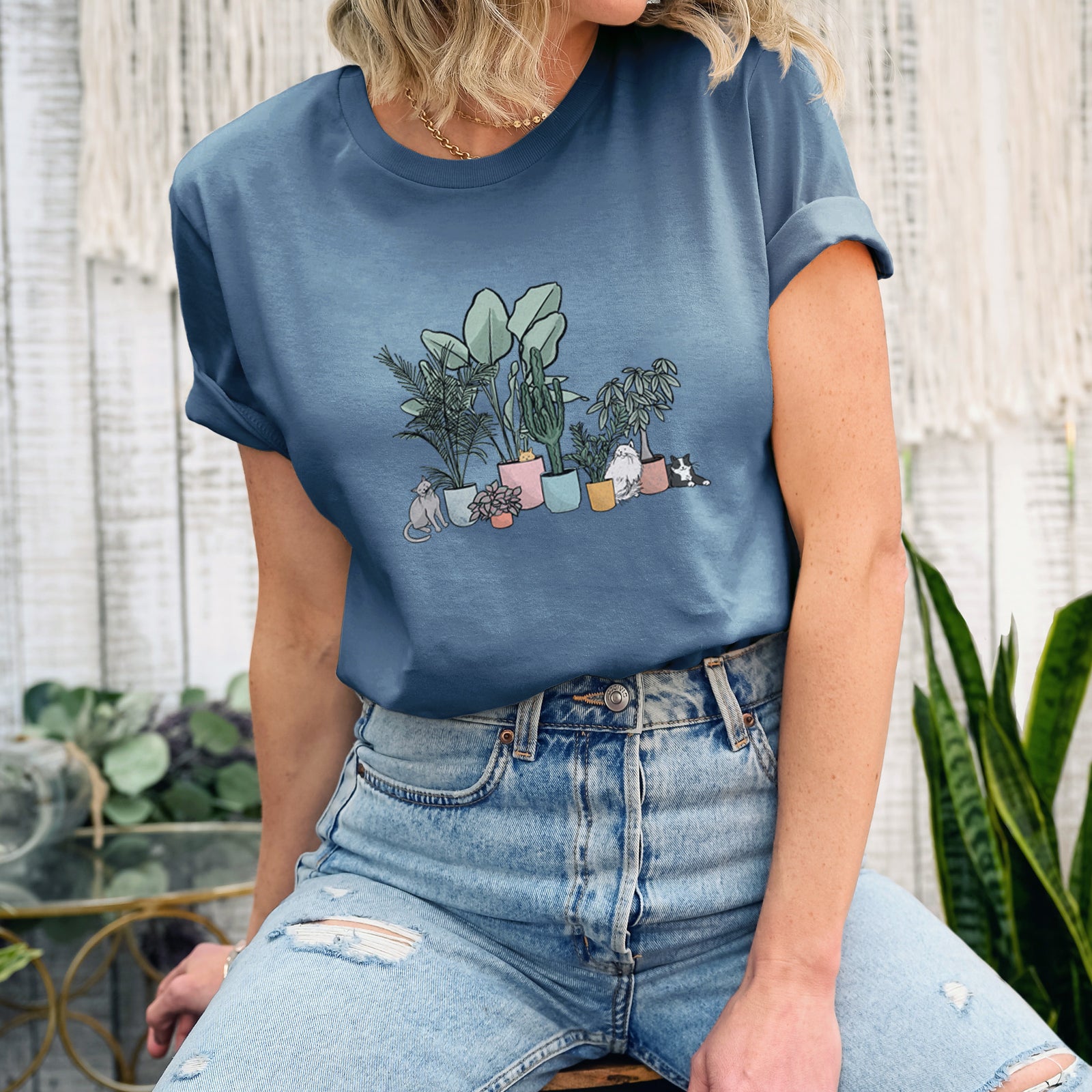 Dusty blue lightweight cotton t-shirt laid flat with the sleeves rolled, printed with a full-color illustration of cats lounging among potted houseplants.