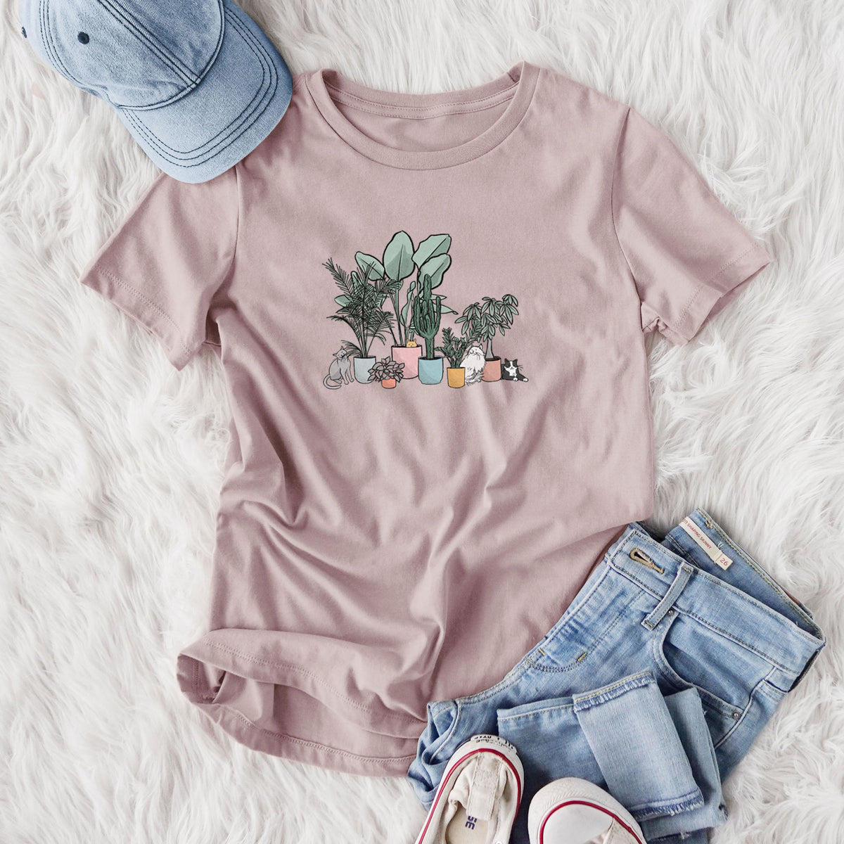 Women's relaxed fit cotton t-shirt printed with a full-color illustration of cats lounging among potted houseplants, styled flat on a white faux fur rug with jeans.