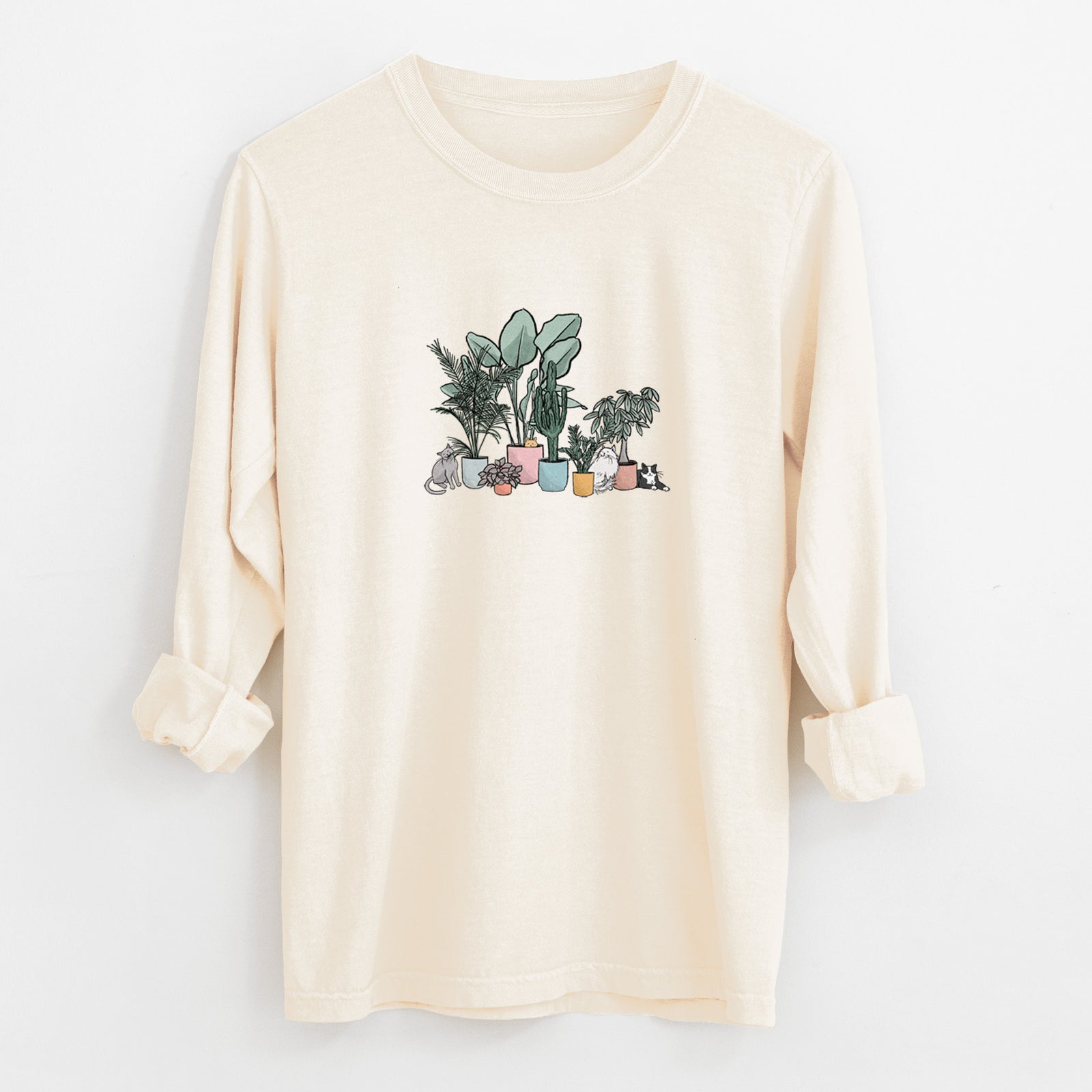 Ivory Comfort Colors long sleeve shirt laid flat with the cuffs rolled, printed with a full-color illustration of cats lounging among potted houseplants.