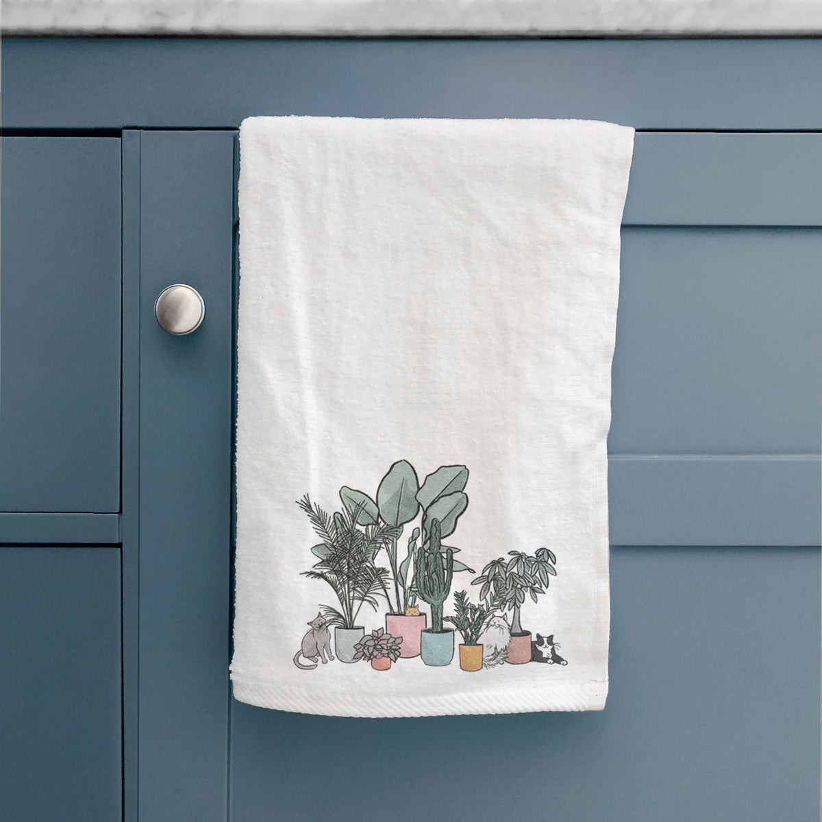White hand towel printed with a full-color illustration of cats lounging among potted houseplants, hanging on a cabinet door.