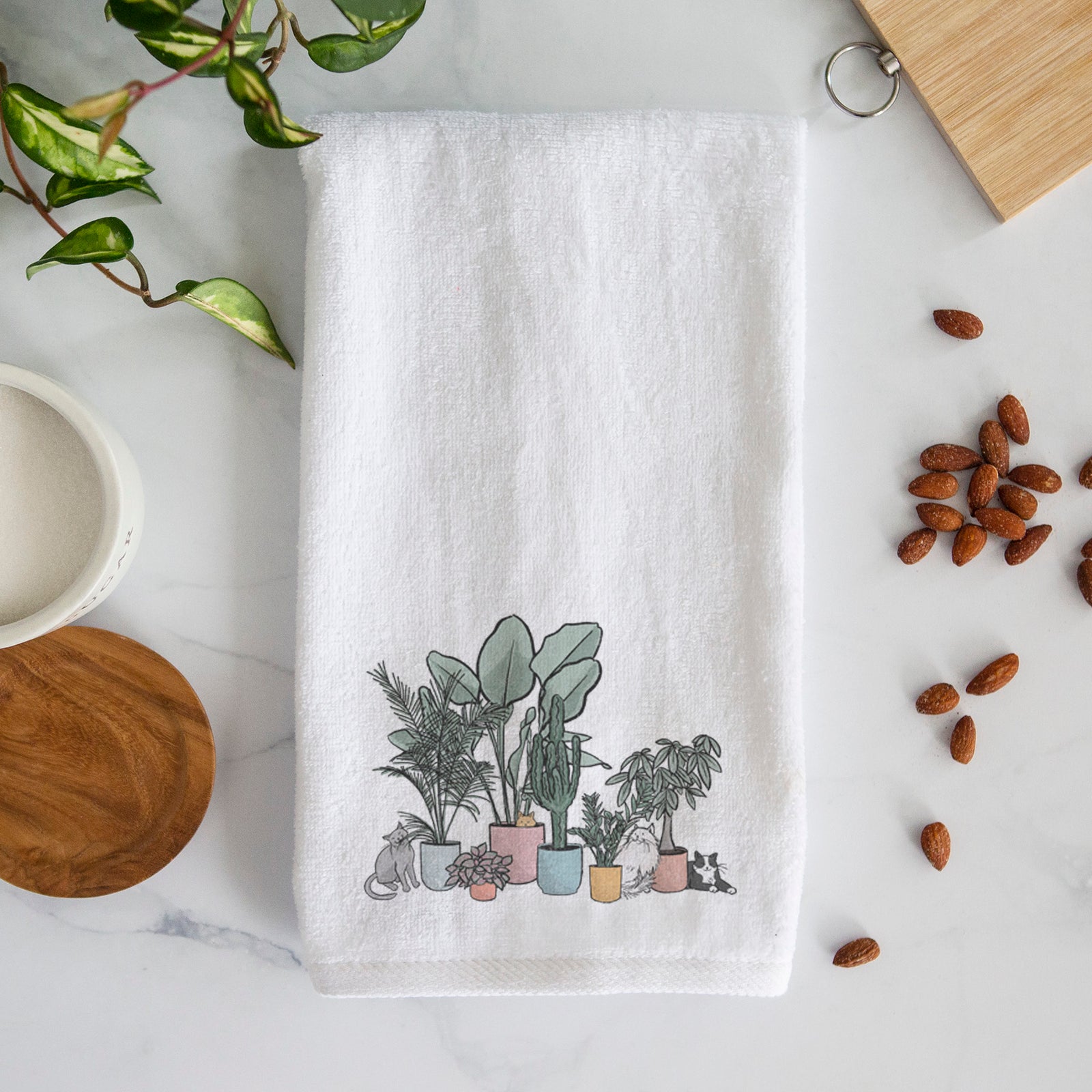 White hand towel printed with a full-color illustration of cats lounging among potted houseplants.