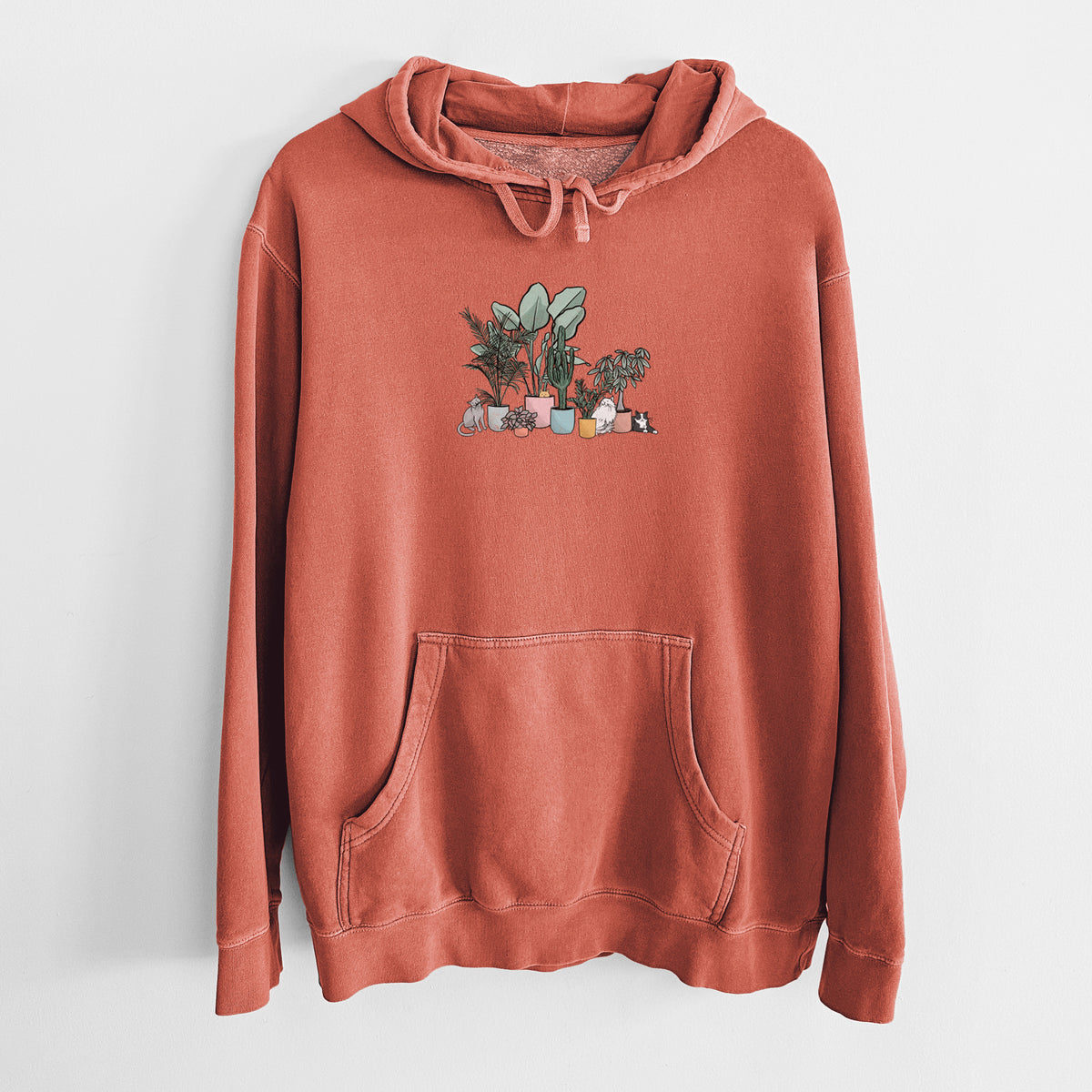 Amber orange pigment dyed hoodie laid flat, printed with a full-color illustration of cats lounging among potted houseplants.