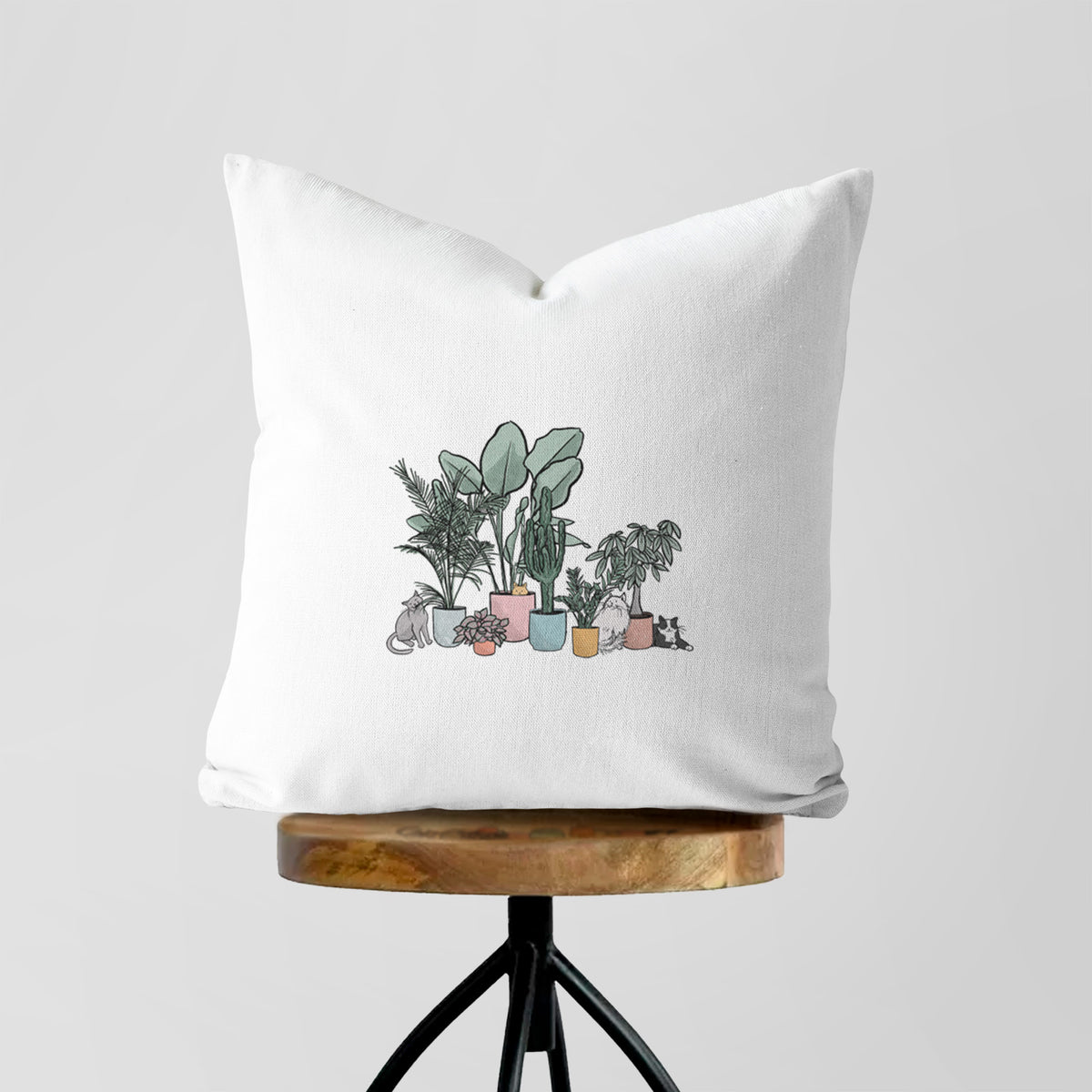 White pillow cover printed with a full-color illustration of cats lounging among potted houseplants, on a wooden stool.