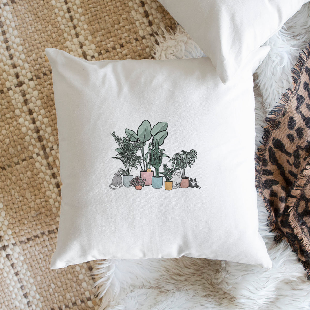 White pillow cover printed with a full-color illustration of cats lounging among potted houseplants, on a woven chair.