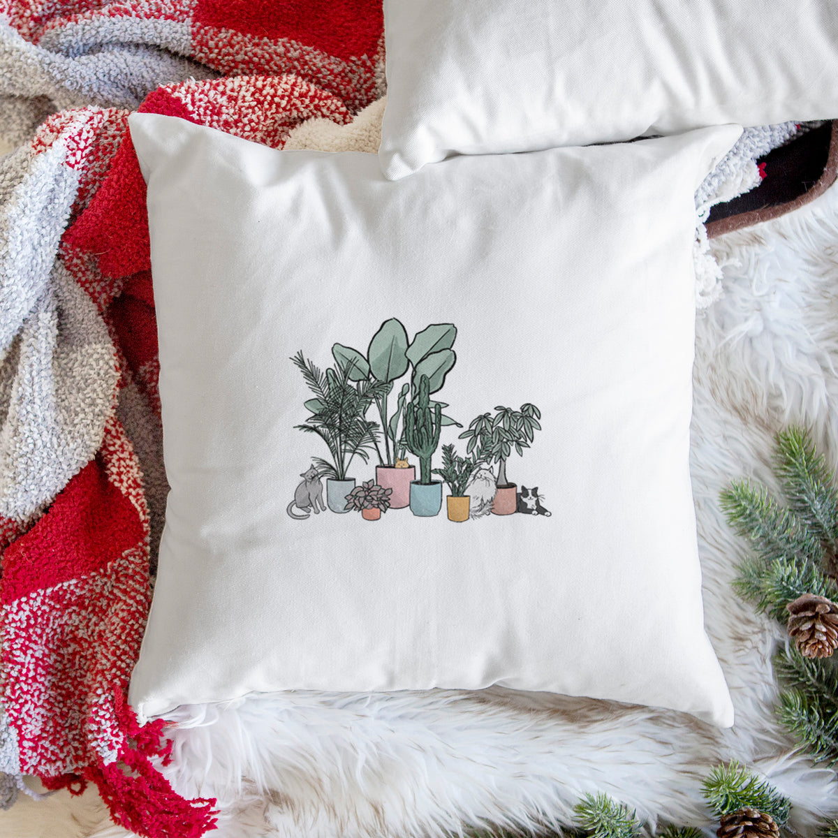 White pillow cover printed with a full-color illustration of cats lounging among potted houseplants, on a woven chair.
