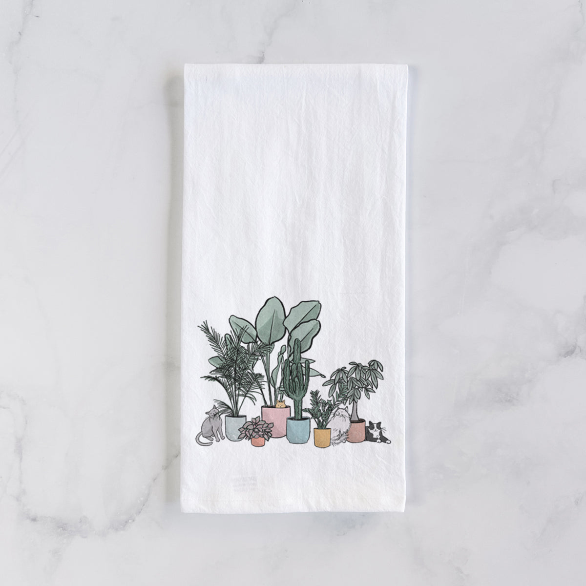 White cotton tea towel laid flat on marble, printed with a full-color illustration of cats lounging among potted houseplants.