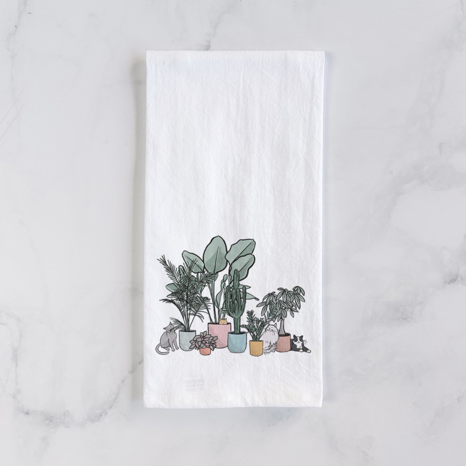 White cotton tea towel printed with a full-color illustration of cats lounging among potted houseplants, on a marble counter beside a round wooden cutting board and greenery.