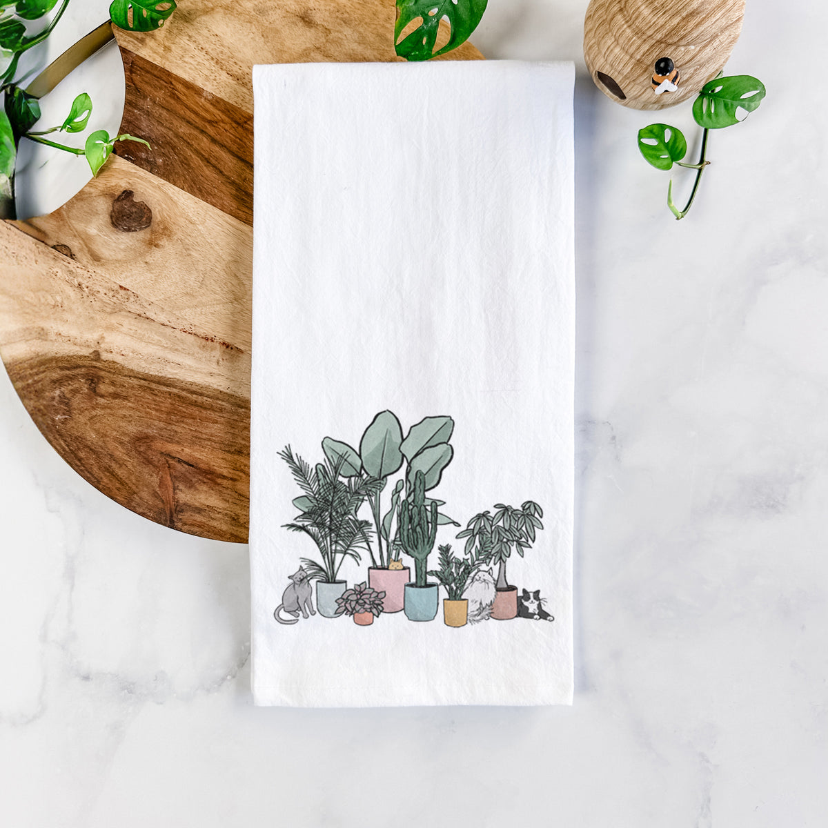 White cotton tea towel printed with a full-color illustration of cats lounging among potted houseplants, on a marble counter beside a round wooden cutting board and greenery.