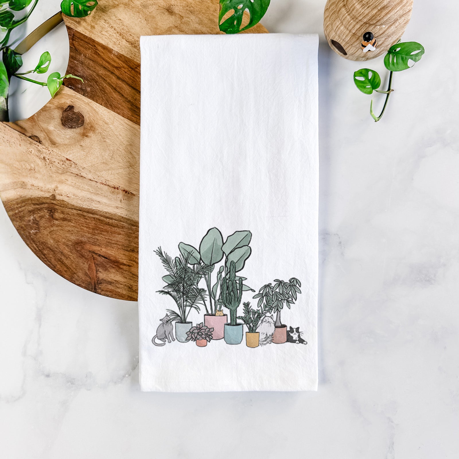 White cotton tea towel printed with a full-color illustration of cats lounging among potted houseplants, on a marble counter beside a round wooden cutting board and greenery.