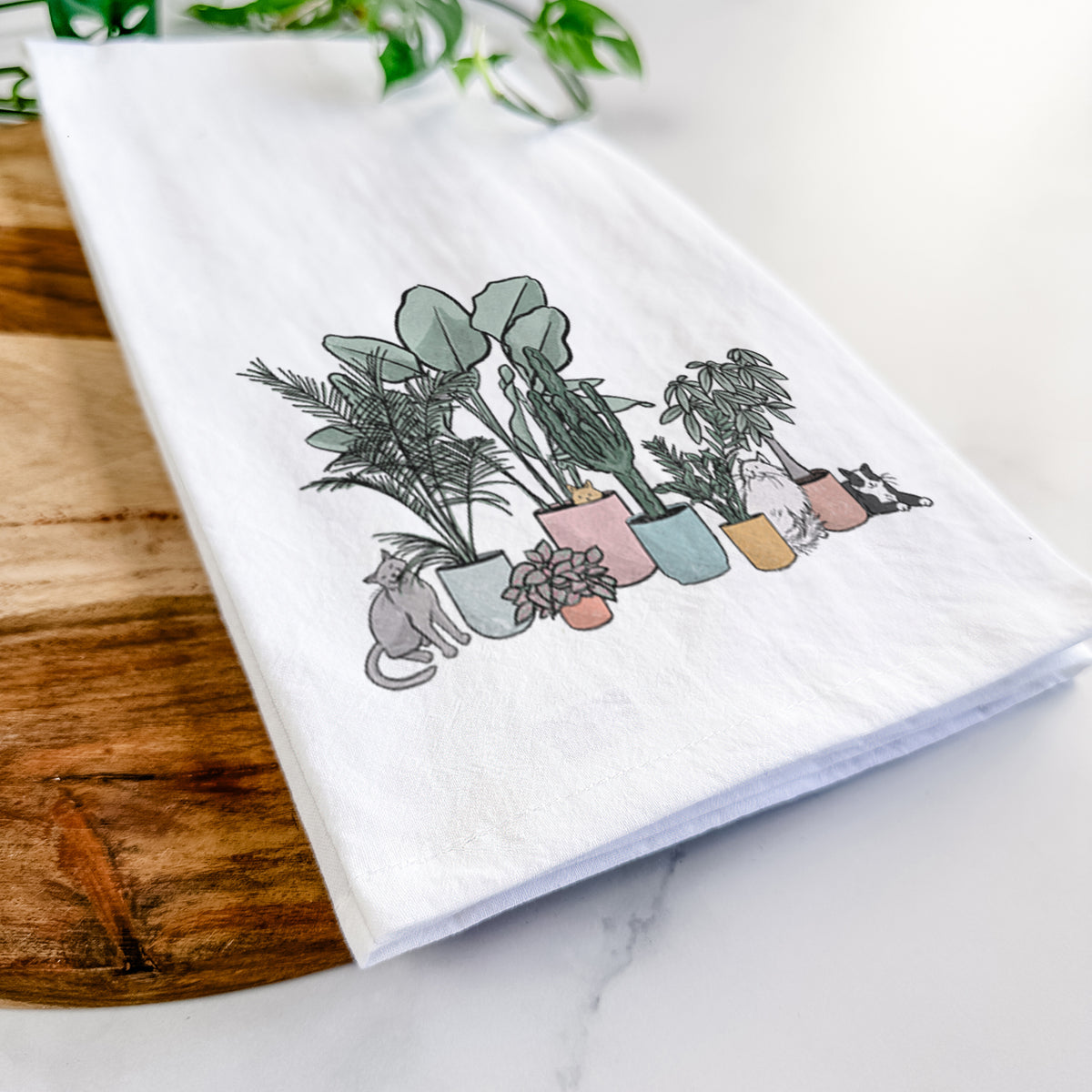 Close-up of a folded white tea towel on a wooden board, showing a full-color illustration of cats lounging among potted houseplants.