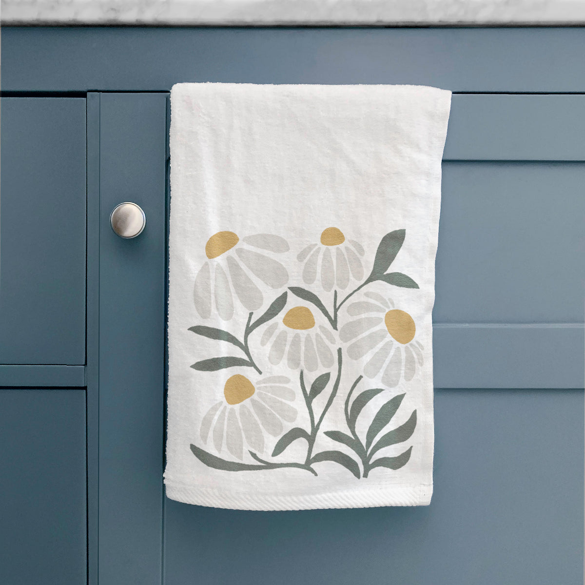 White hand towel printed with a soft full-color illustration of white chamomile flowers and green leaves, hanging on a cabinet door.