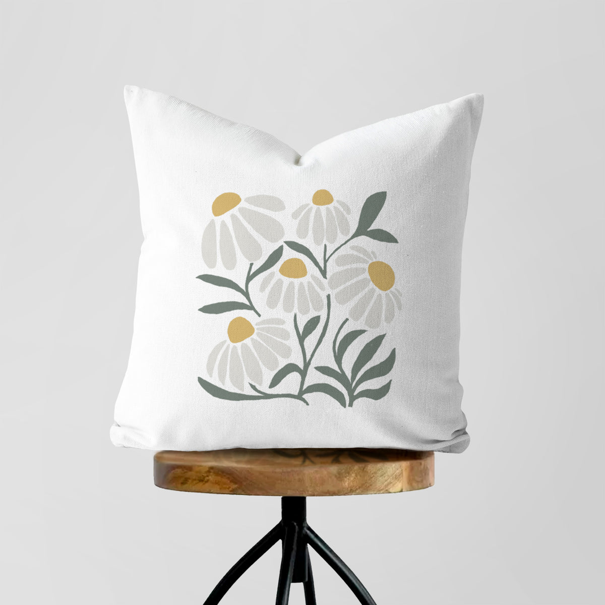 White pillow cover printed with a soft full-color illustration of white chamomile flowers and green leaves, on a wooden stool.