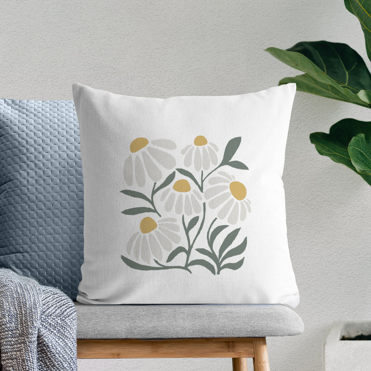 White pillow cover printed with a soft full-color illustration of white chamomile flowers and green leaves, on a bench beside plants.