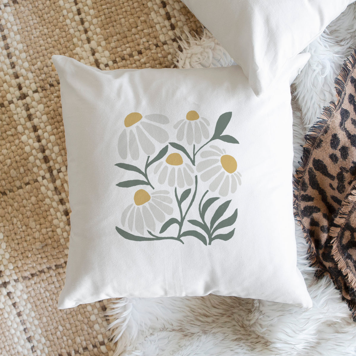 White pillow cover printed with a soft full-color illustration of white chamomile flowers and green leaves, on a woven chair.