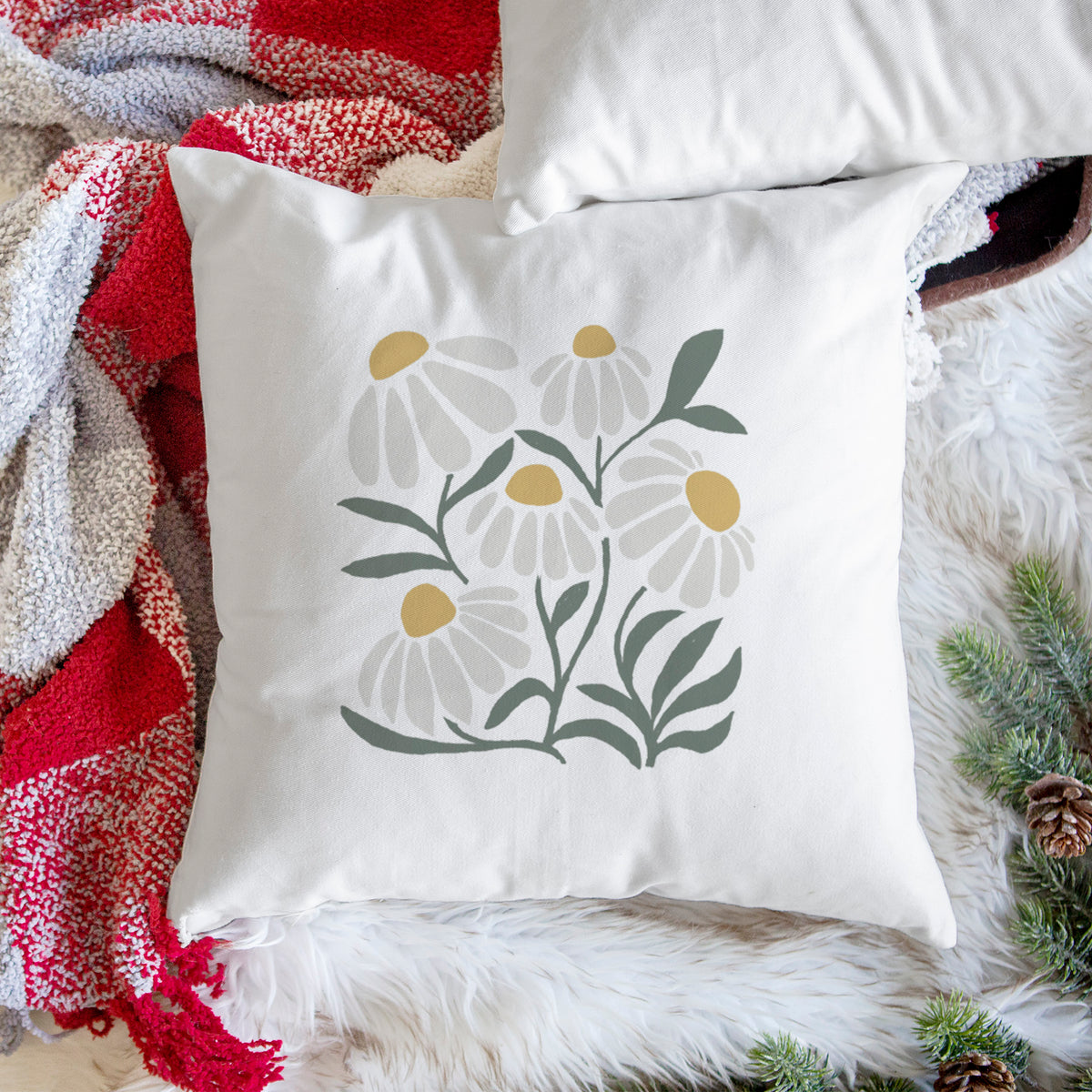 White pillow cover printed with a soft full-color illustration of white chamomile flowers and green leaves, on a woven chair.