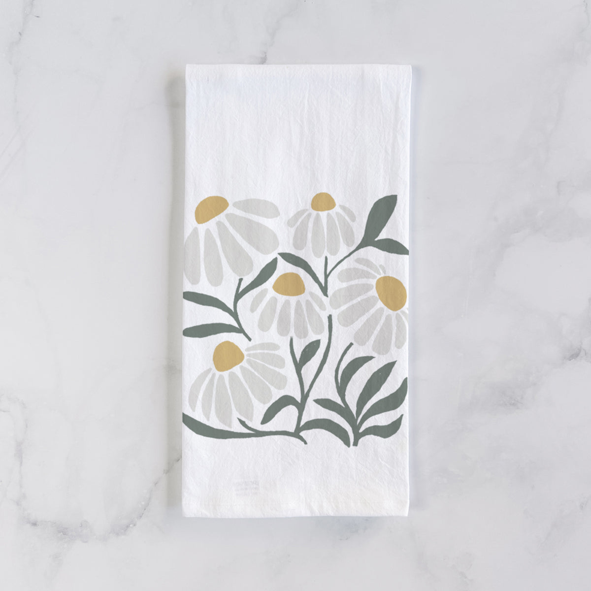 White cotton tea towel laid flat on marble, printed with a soft full-color illustration of white chamomile flowers and green leaves.