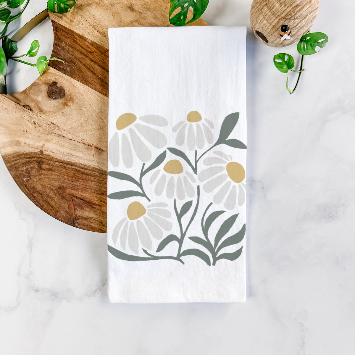 White cotton tea towel printed with a soft full-color illustration of white chamomile flowers and green leaves, on a marble counter beside a round wooden cutting board and greenery.