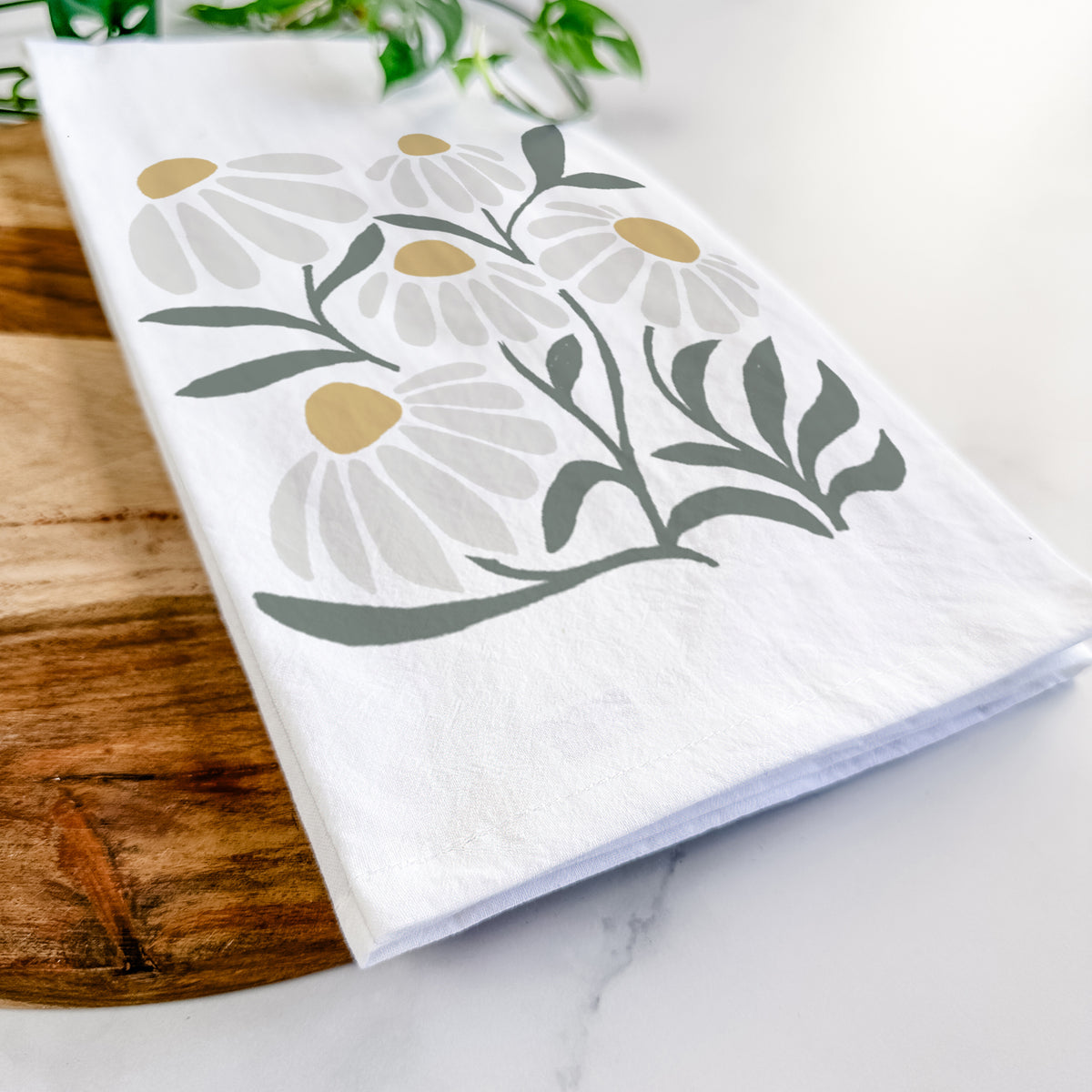 Close-up of a folded white tea towel on a wooden board, showing a soft full-color illustration of white chamomile flowers and green leaves.