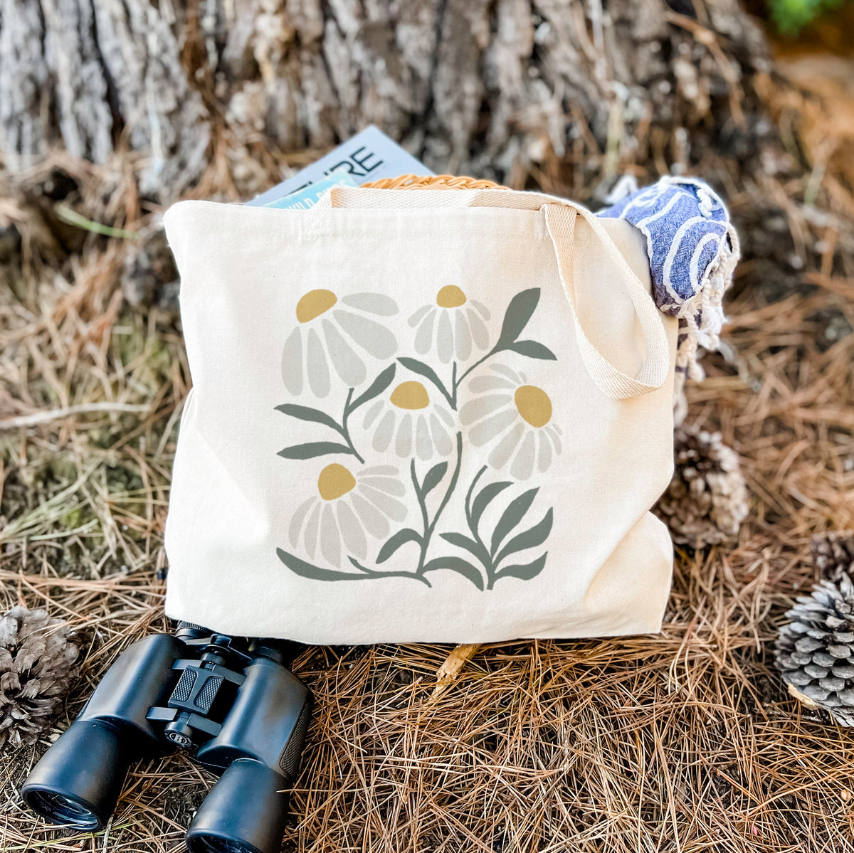 Canvas tote bag printed with a soft full-color illustration of white chamomile flowers and green leaves, resting on dry grass beside a pair of binoculars.
