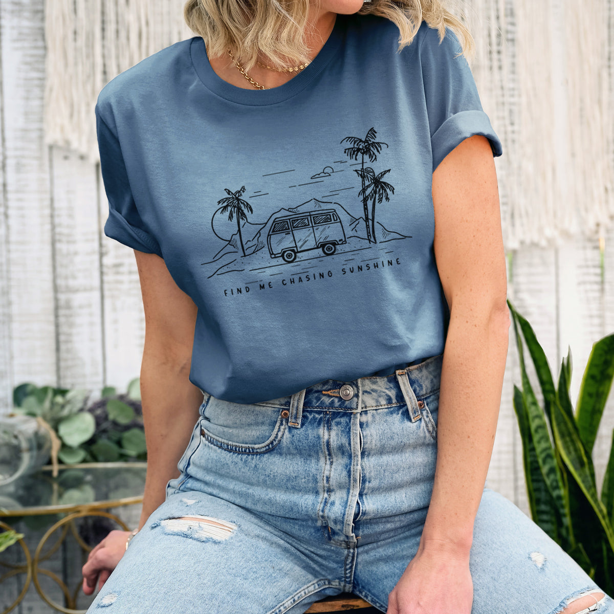 Woman wearing a steel blue lightweight cotton t-shirt printed with a black ink illustration of a camper van under palm trees and the sun, with the words Find Me Chasing Sunshine.