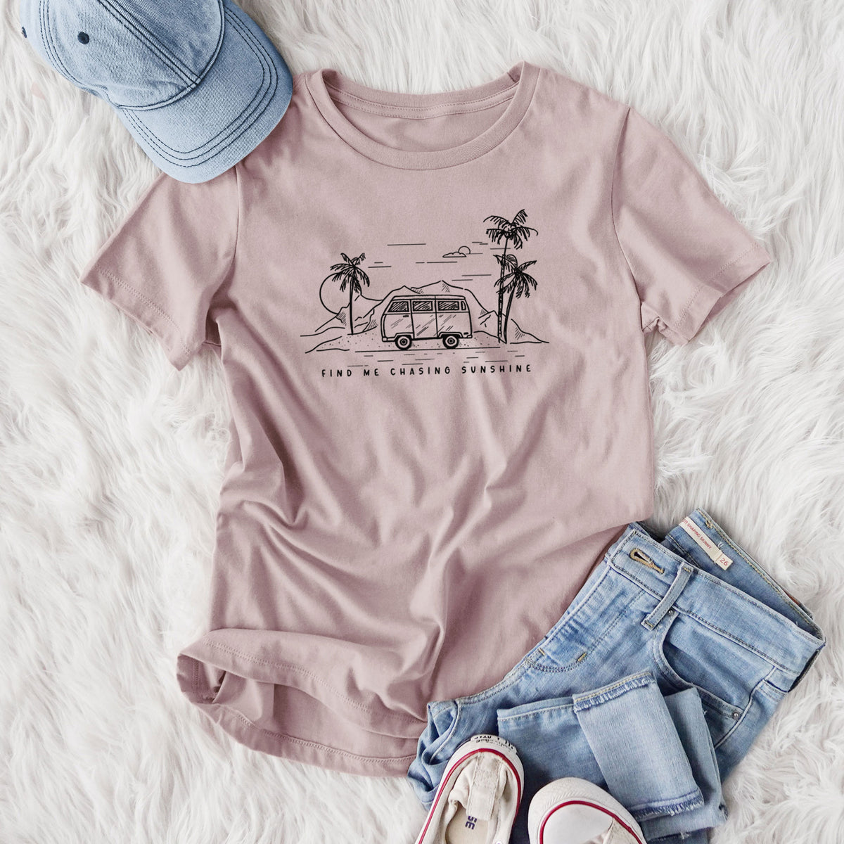 Women's relaxed fit cotton t-shirt printed with a black ink illustration of a camper van under palm trees and the sun, with the words Find Me Chasing Sunshine, styled flat on a white faux fur rug with jeans.