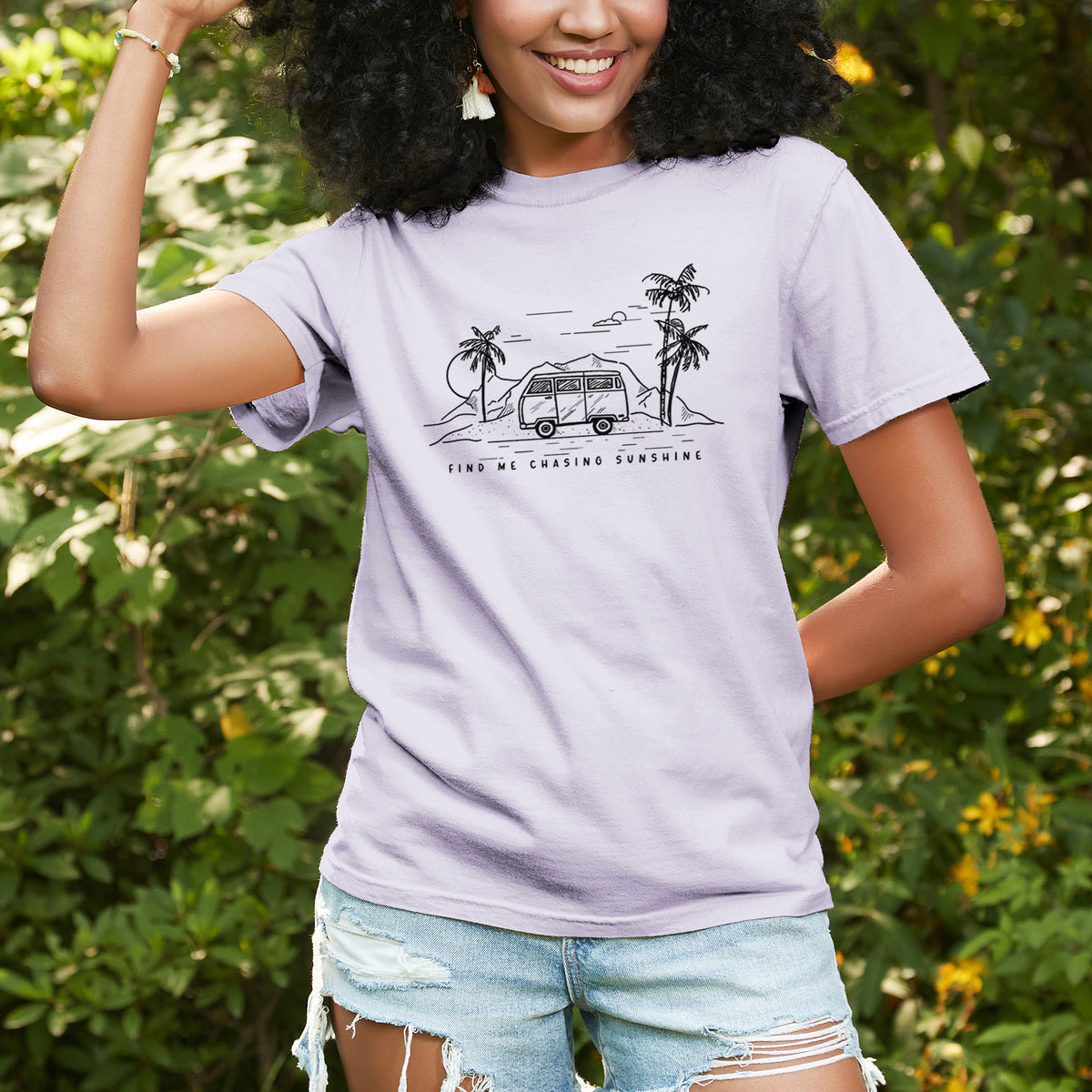 Woman wearing a Comfort Colors t-shirt printed with a black ink illustration of a camper van under palm trees and the sun, with the words Find Me Chasing Sunshine, outdoors.