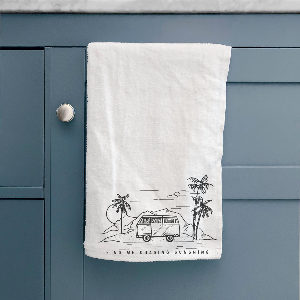 White hand towel printed with a black ink illustration of a camper van under palm trees and the sun, with the words Find Me Chasing Sunshine, hanging on a cabinet door.