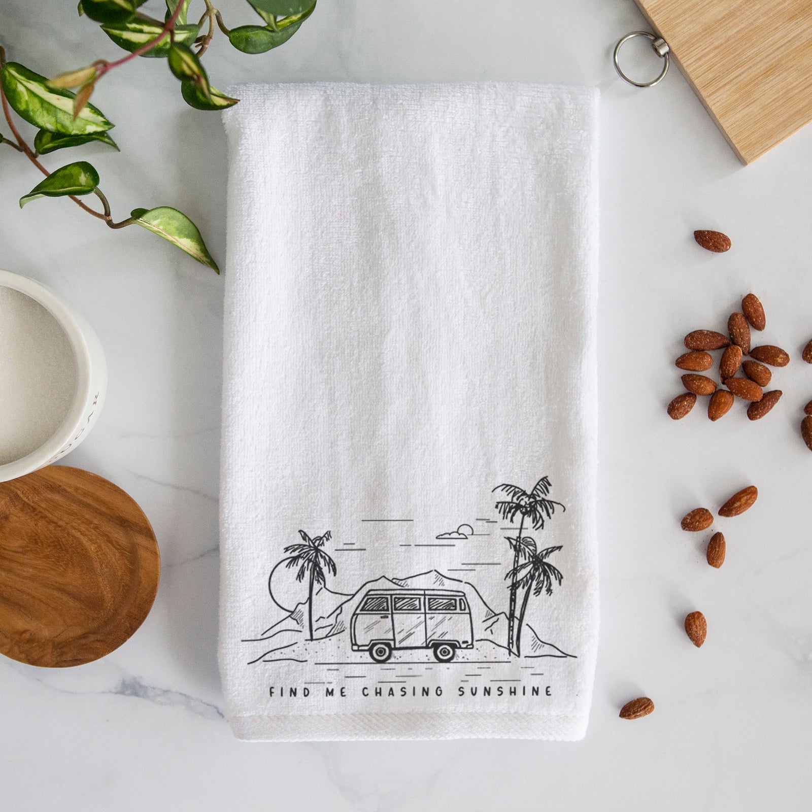 White hand towel printed with a black ink illustration of a camper van under palm trees and the sun, with the words Find Me Chasing Sunshine.
