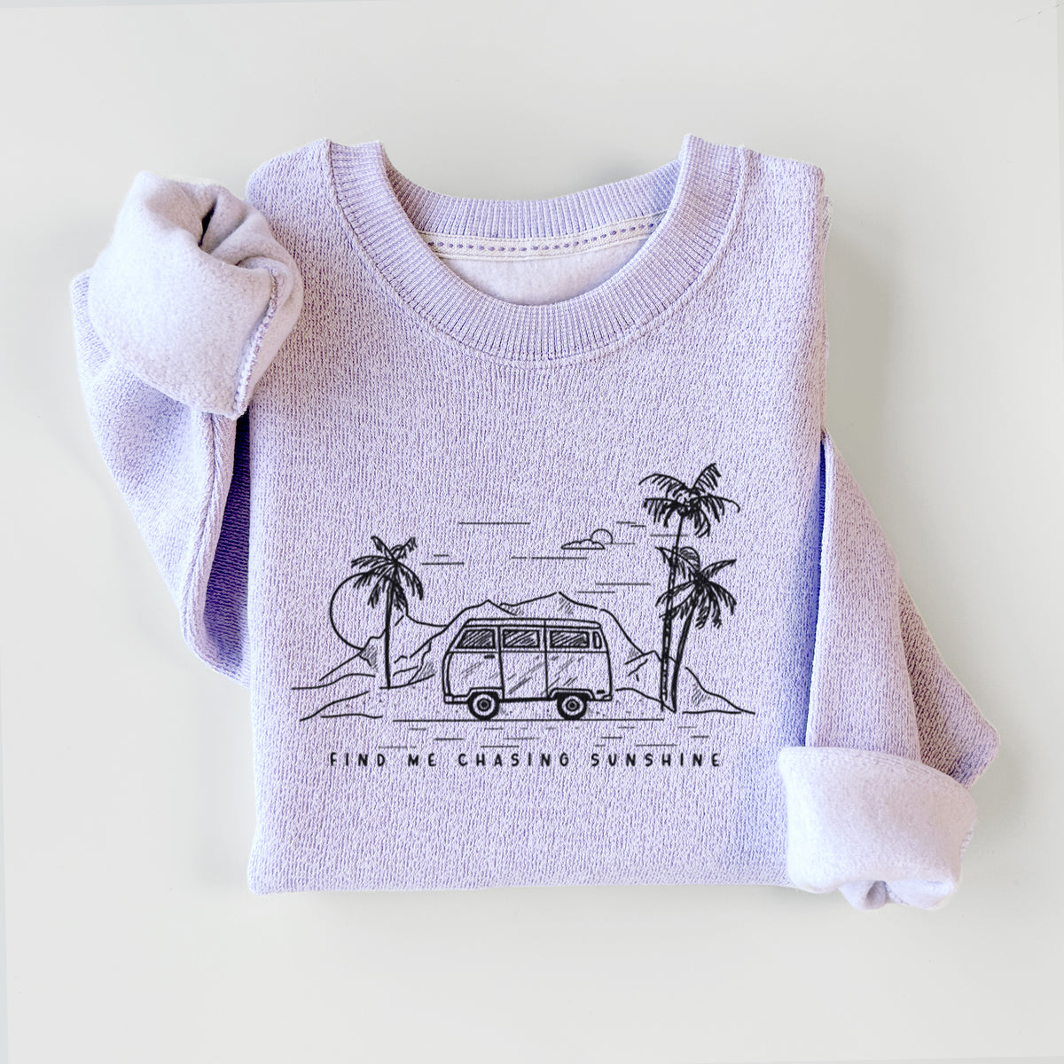 Ice purple knit sweatshirt printed with a black ink illustration of a camper van under palm trees and the sun, with the words Find Me Chasing Sunshine, styled flat.