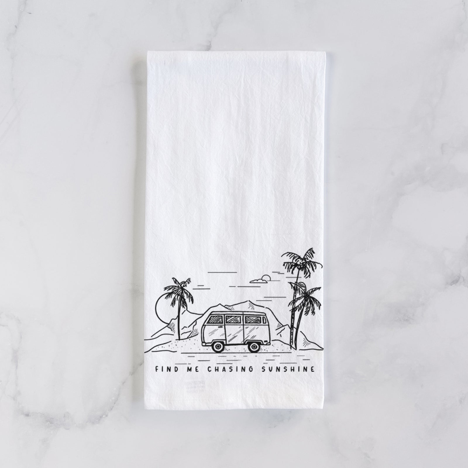 White cotton tea towel printed with a black ink illustration of a camper van under palm trees and the sun, with the words Find Me Chasing Sunshine, on a marble counter beside a round wooden cutting board and greenery.