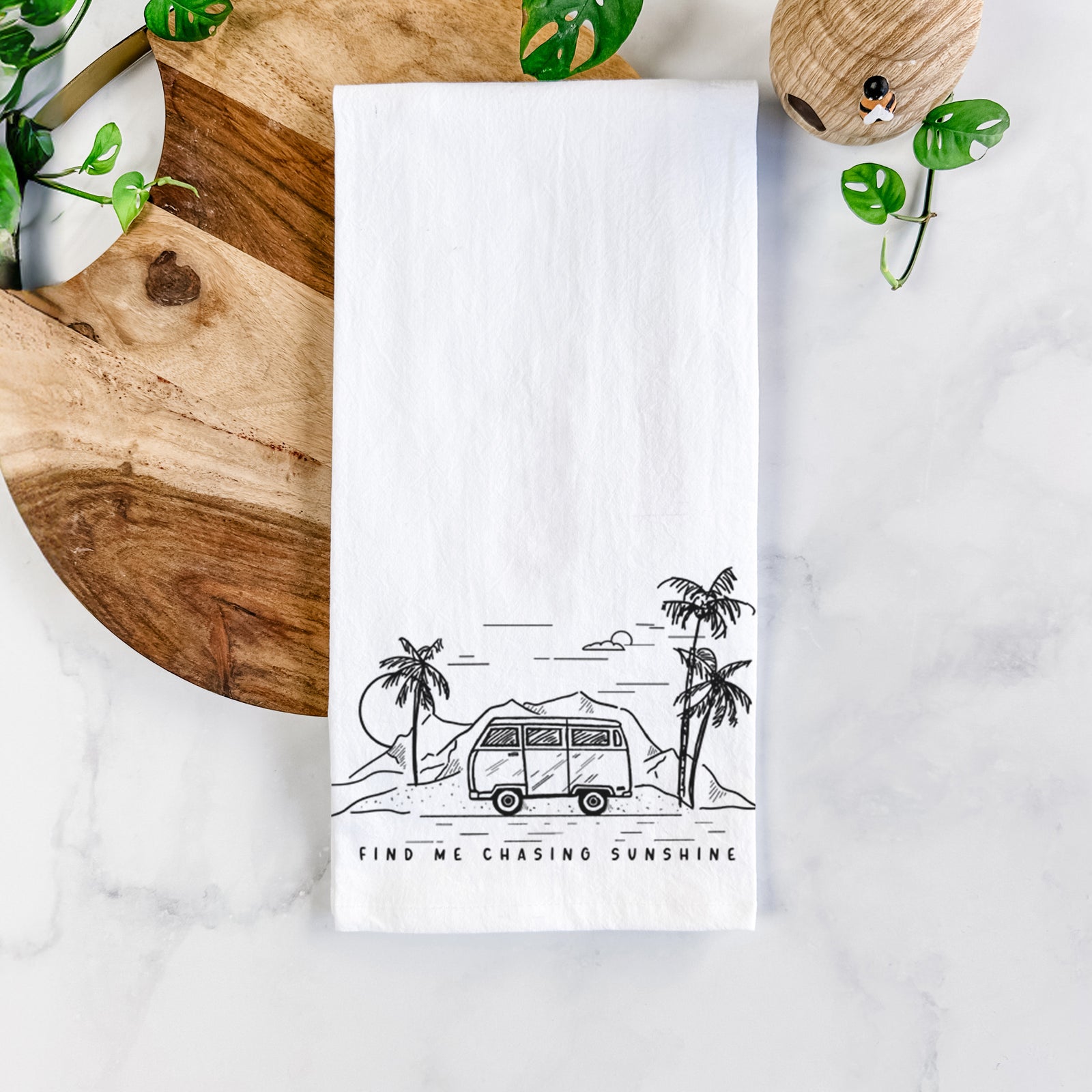 White cotton tea towel printed with a black ink illustration of a camper van under palm trees and the sun, with the words Find Me Chasing Sunshine, on a marble counter beside a round wooden cutting board and greenery.