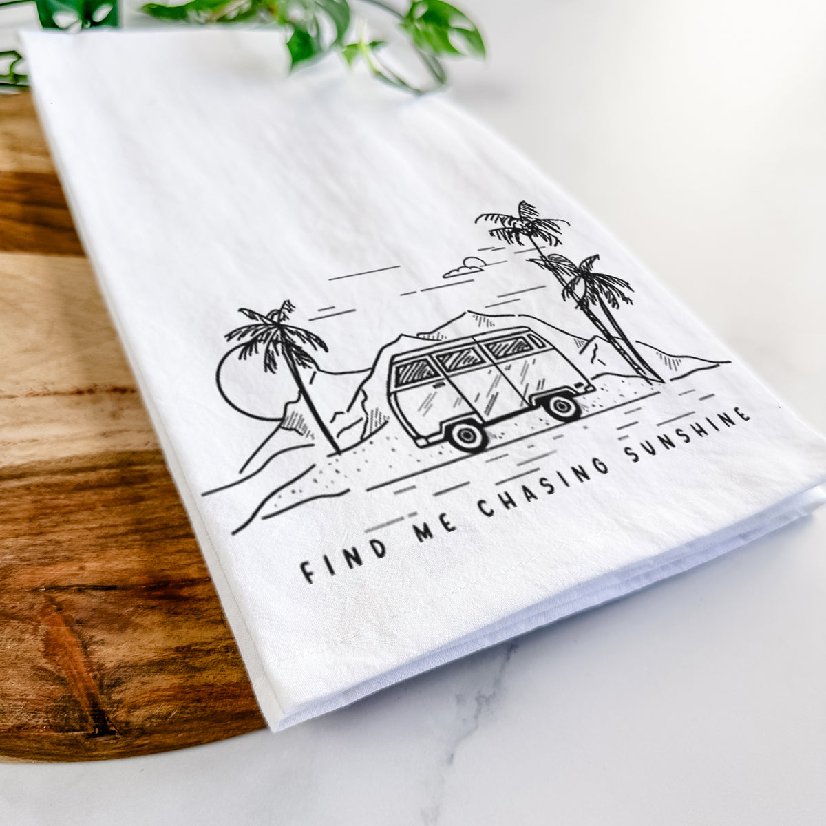 Close-up of a folded white tea towel on a wooden board, showing a black ink illustration of a camper van under palm trees and the sun, with the words Find Me Chasing Sunshine.