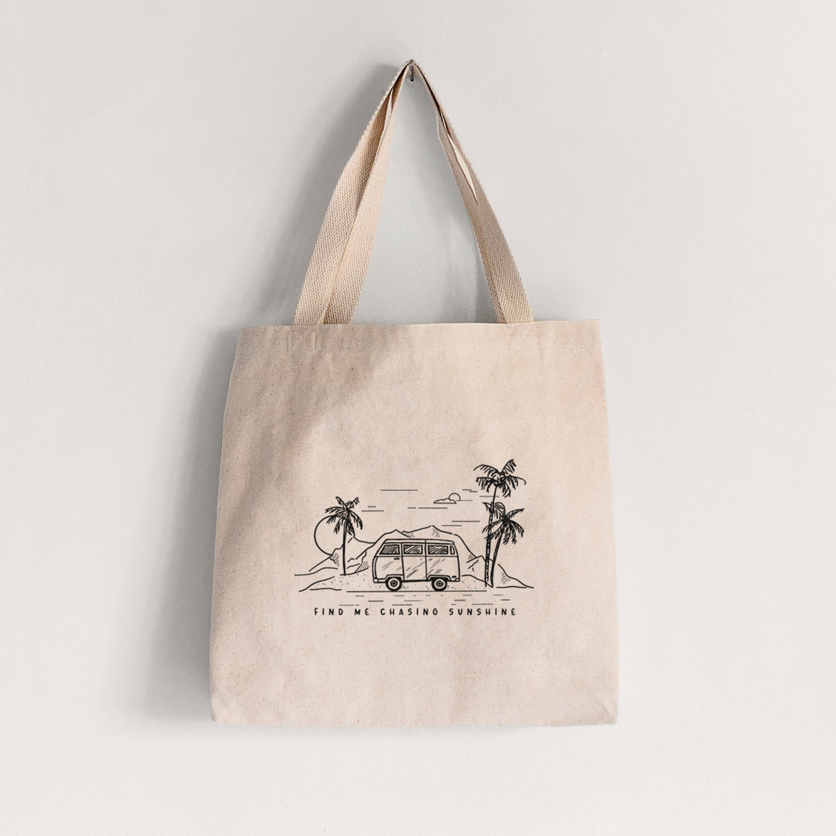 Natural canvas tote bag printed with a black ink illustration of a camper van under palm trees and the sun, with the words Find Me Chasing Sunshine.