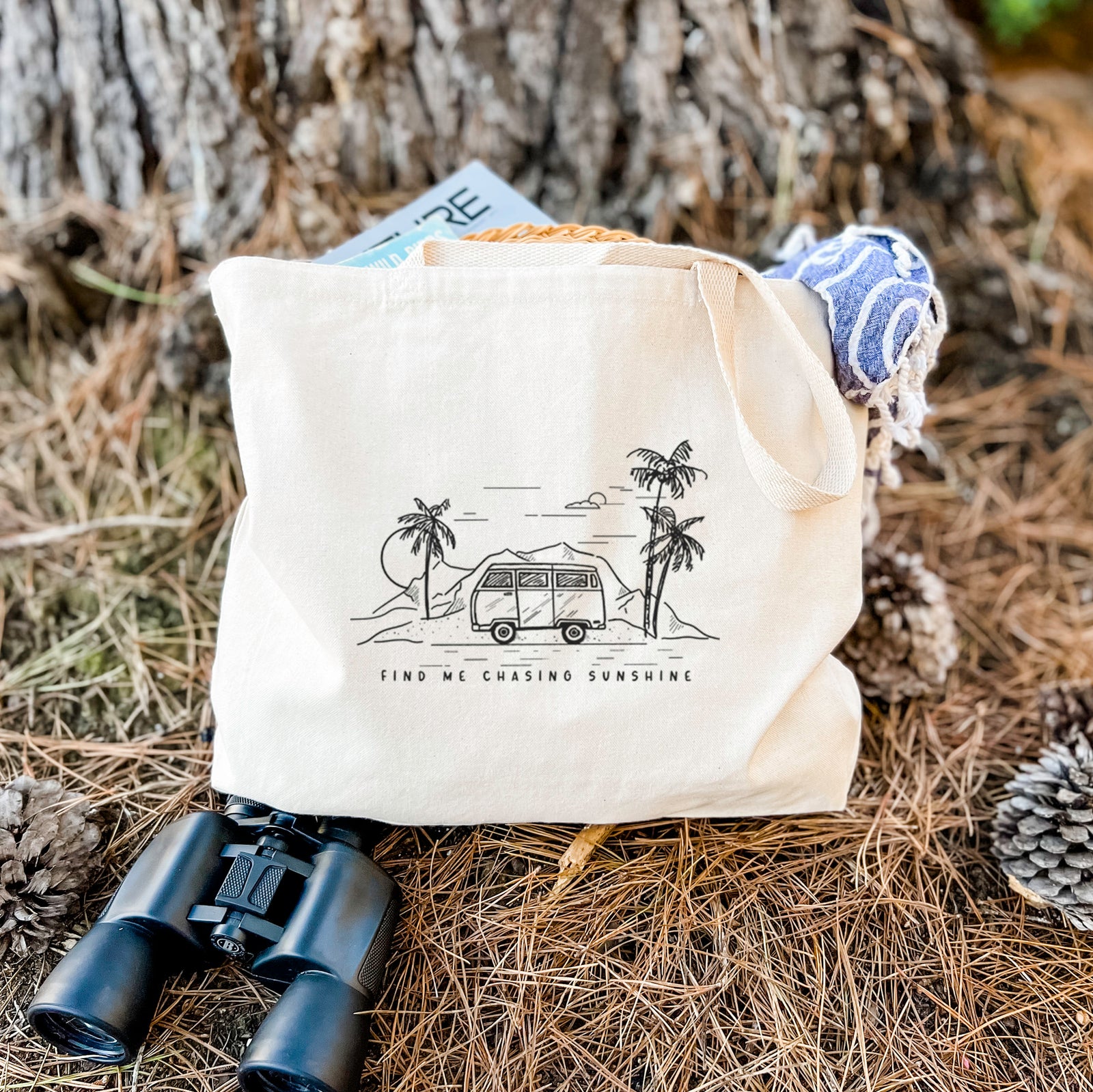 Natural canvas tote bag printed with a black ink illustration of a camper van under palm trees and the sun, with the words Find Me Chasing Sunshine.