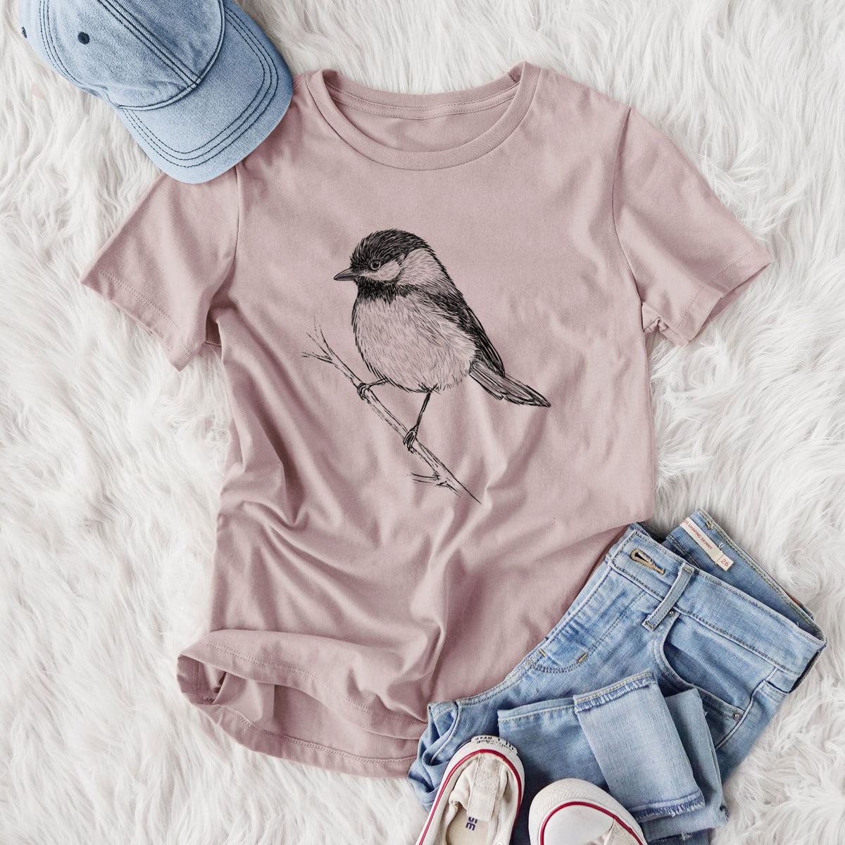 Women's relaxed fit cotton t-shirt printed with a detailed black ink sketch of a black-capped chickadee perched on a branch, styled flat on a white faux fur rug with jeans.