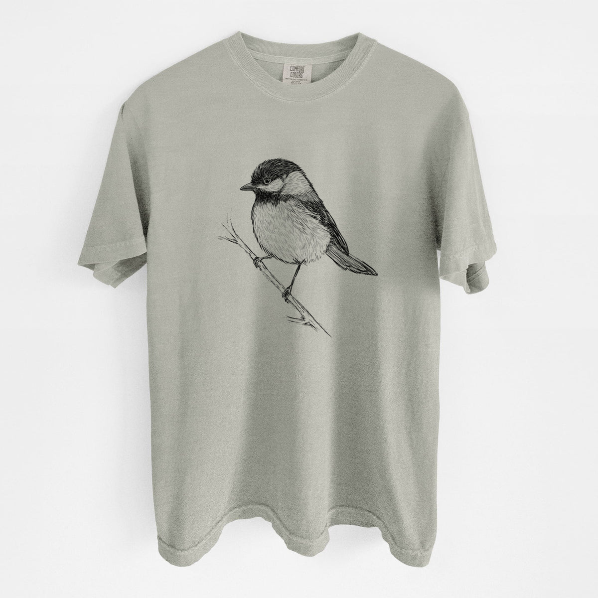 Sandstone beige Comfort Colors t-shirt laid flat, printed with a detailed black ink sketch of a black-capped chickadee perched on a branch.